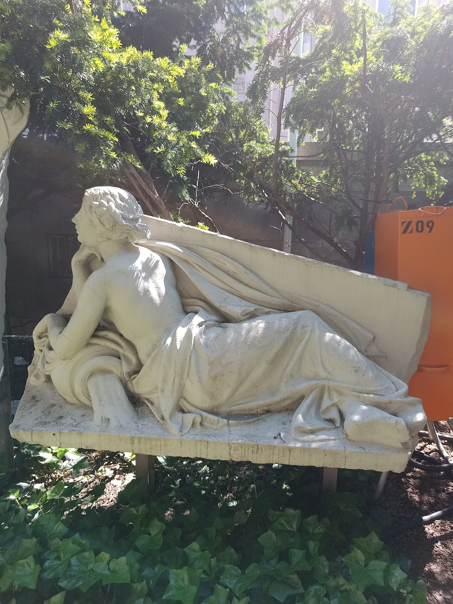 Here is a detailed description of the image:
**General Overview:** The image depicts a white sculpture of a reclining figure, possibly female, placed outdoors in what appears to be a park or garden setting. The area is shaded by trees, and there's an orange container labeled "Z09" in the background.
**Detailed Description:**
* **Sculpture:** The sculpture is made of a white material, likely stone or a similar substance, and depicts a figure lying on a flat surface. The figure is semi-draped in what seems to be fabric. The head rests on the left hand, suggesting a pose of contemplation or rest. The sculpture shows signs of weathering.
* **Surroundings:** The sculpture is surrounded by greenery, specifically a patch of green ivy-like plants at its base. The background includes trees and a structure with window-like openings. The orange container is adjacent to the sculpture on the right side.
**Location:** The scene appears to be located in an urban park or garden. The presence of the orange container suggests ongoing maintenance or construction in the area.
**People:** There are no people visible in the image, only the sculpture.