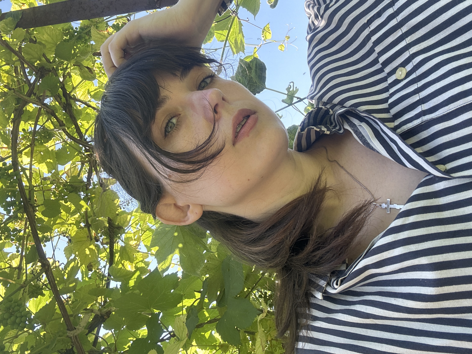 A low-angle, close-up selfie captures a young woman with dark brown hair and light green eyes, positioned in the foreground. She is outdoors, with lush green grape leaves and unripe green grapes arching over her head, set against a bright blue sky. She wears a black and white striped button-down shirt and a silver cross necklace. Her right hand is raised, resting on her forehead, and her gaze is directed slightly upward and to the left of the camera. The image appears to be taken in Zvonarevka, Russia, as indicated by the presence of grapevines, a common agricultural feature in the region.