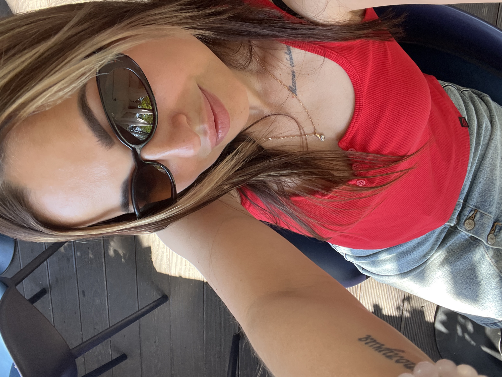 A woman with long, brown hair with blonde highlights, wearing sunglasses, a red tank top, and a denim skirt, is taking a selfie. She has a necklace with a small pendant and a tattoo on her chest. Another tattoo is visible on her arm. She is seated on a dark blue chair, with a wooden floor visible behind her. The scene is outdoors, likely in a cafe or restaurant setting, with shadows cast by sunlight.