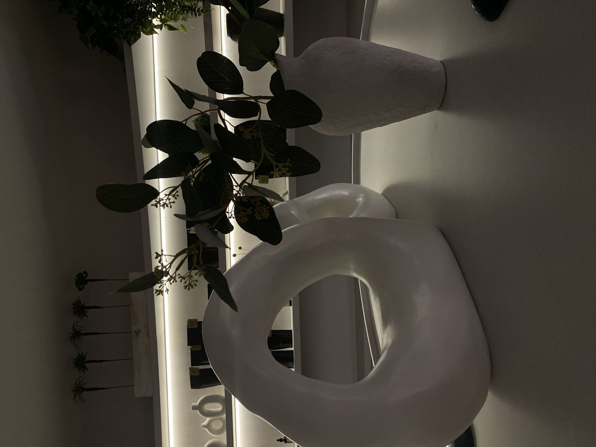 The image shows a modern interior with a focus on minimalist decor. Two white vases are prominently displayed. One is a rounded, textured vase, and the other is a large, circular, donut-shaped sculpture. Green foliage is arranged in the vases, adding a natural element. The background reveals a shelf with various decorative items and illuminated vertical panels. The scene is well-lit and has a contemporary aesthetic. It is impossible to definitively pinpoint the location as Votkinsk, Russia, without additional verifiable information tying the decor or specific identifiers to that city.