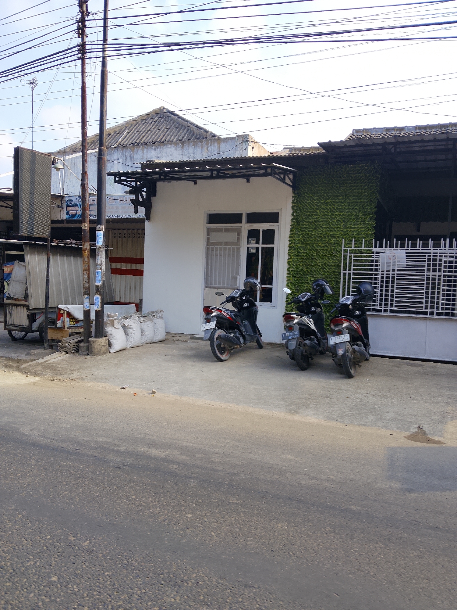 In the city of Ngelo, Indonesia, a street scene unfolds with a white building featuring a green, textured facade resembling stacked leaves. In front of the building, three black motorcycles are parked on a concrete surface. The motorcycle on the far left is partially obscured by the building's corner. The middle motorcycle is facing right, and the motorcycle on the far right is also facing right, positioned slightly behind the middle one. All three motorcycles have riders wearing black helmets. To the left of the motorcycles, a small cart with a silver canopy is visible, positioned near a utility pole. Several large white sacks are stacked on the ground to the left of the building. The environment suggests a commercial or residential area with a paved road in the foreground.