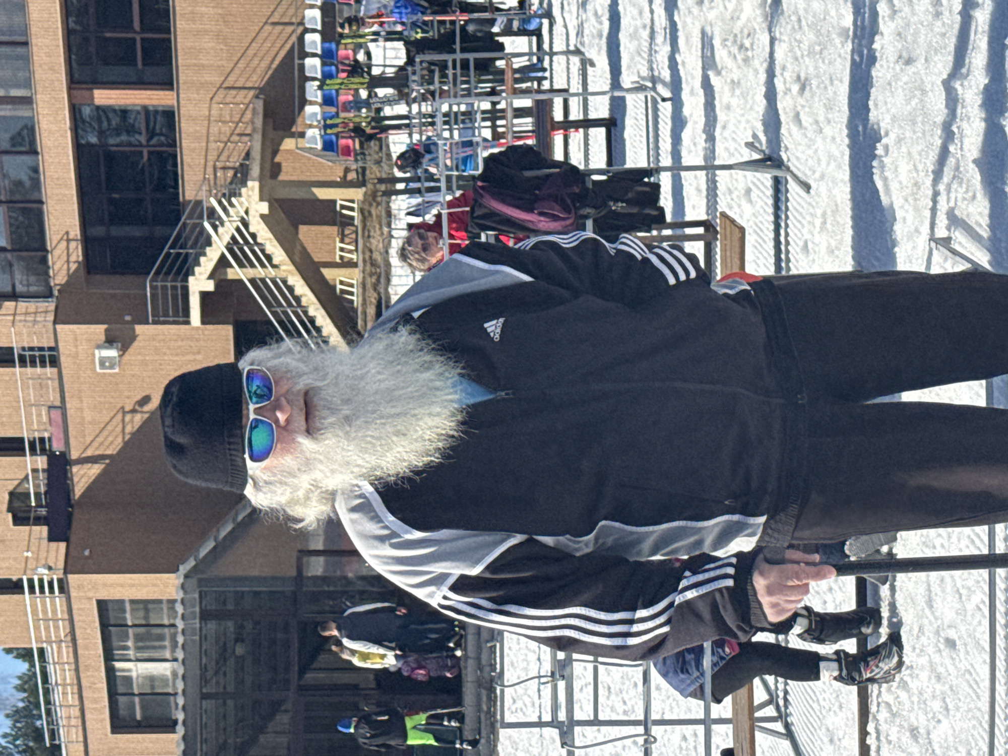 The image depicts a man with a long, gray beard wearing a black beanie and sunglasses, sitting in a ski lift. He is wearing a black Adidas jacket with white stripes. In the background, there is a building, snow, and more ski lift chairs. The location is likely Прикамский, Russia, a city known for its ski resorts. Other people are visible in the background, both sitting on and standing near the ski lift. The man appears to be riding the ski lift upwards.