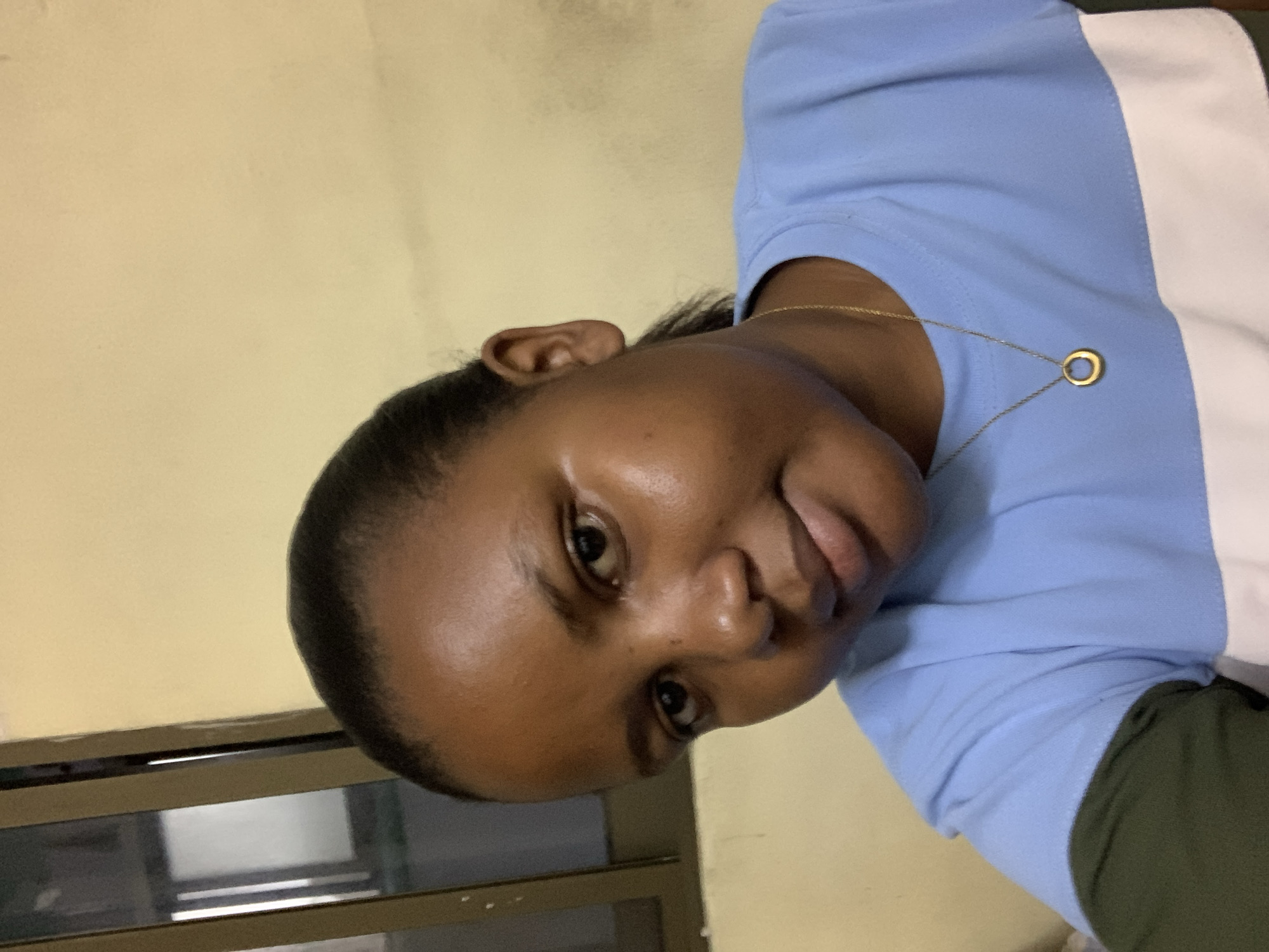 A medium shot depicts a woman with dark skin and short, dark hair, wearing a light blue and white shirt, and a gold necklace with a circular pendant. She is facing the camera with a slight smile. Behind her is a cream-colored wall and what appears to be a window or cabinet with a dark frame. The location in Dar es-Salaam, Tanzania is not discernible from this image.