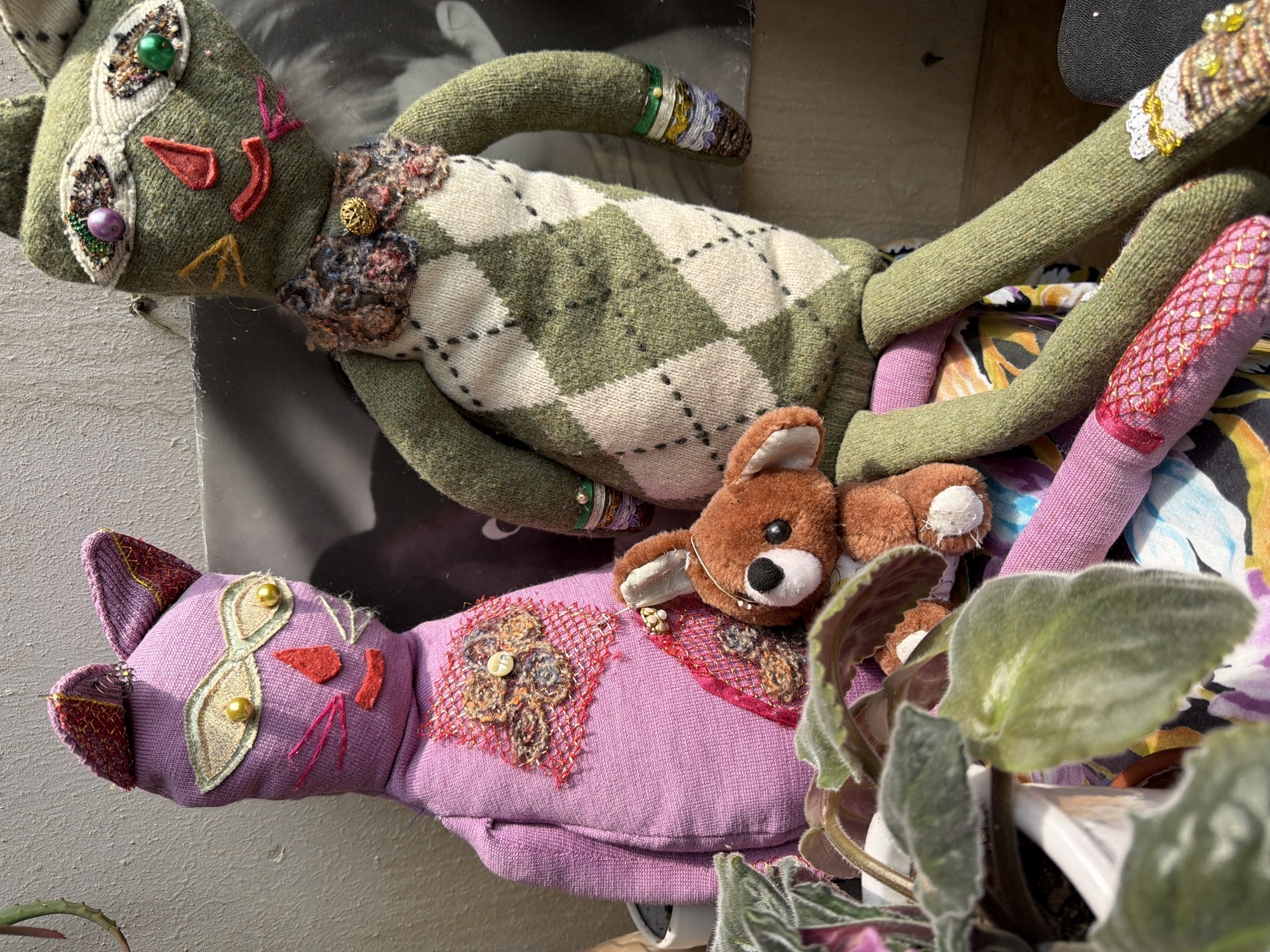 Two handmade fabric dolls, one purple cat and one green cat, are seated side-by-side. The purple cat has large, gold-bead eyes, red fabric lips and triangle nose, and embroidered whiskers. Its chest is adorned with a decorative patch of woven fibers and a small white button. The green cat has multiple bead eyes in various colors, red fabric triangle ears, and a red fabric mouth. It wears a knitted argyle sweater and a ruffled collar with a gold bead accent. Both cats have visible stitching and embellishments on their arms and legs. A small, brown, plush bear is positioned between the two cats. In the foreground, the leafy green foliage of a potted plant is visible. The background consists of a textured gray wall and a large, black-and-white photographic print. The scene appears to be an indoor setting, possibly a home, with natural light casting shadows. There is no indication of the city of Izhevsk, Russia in the image.