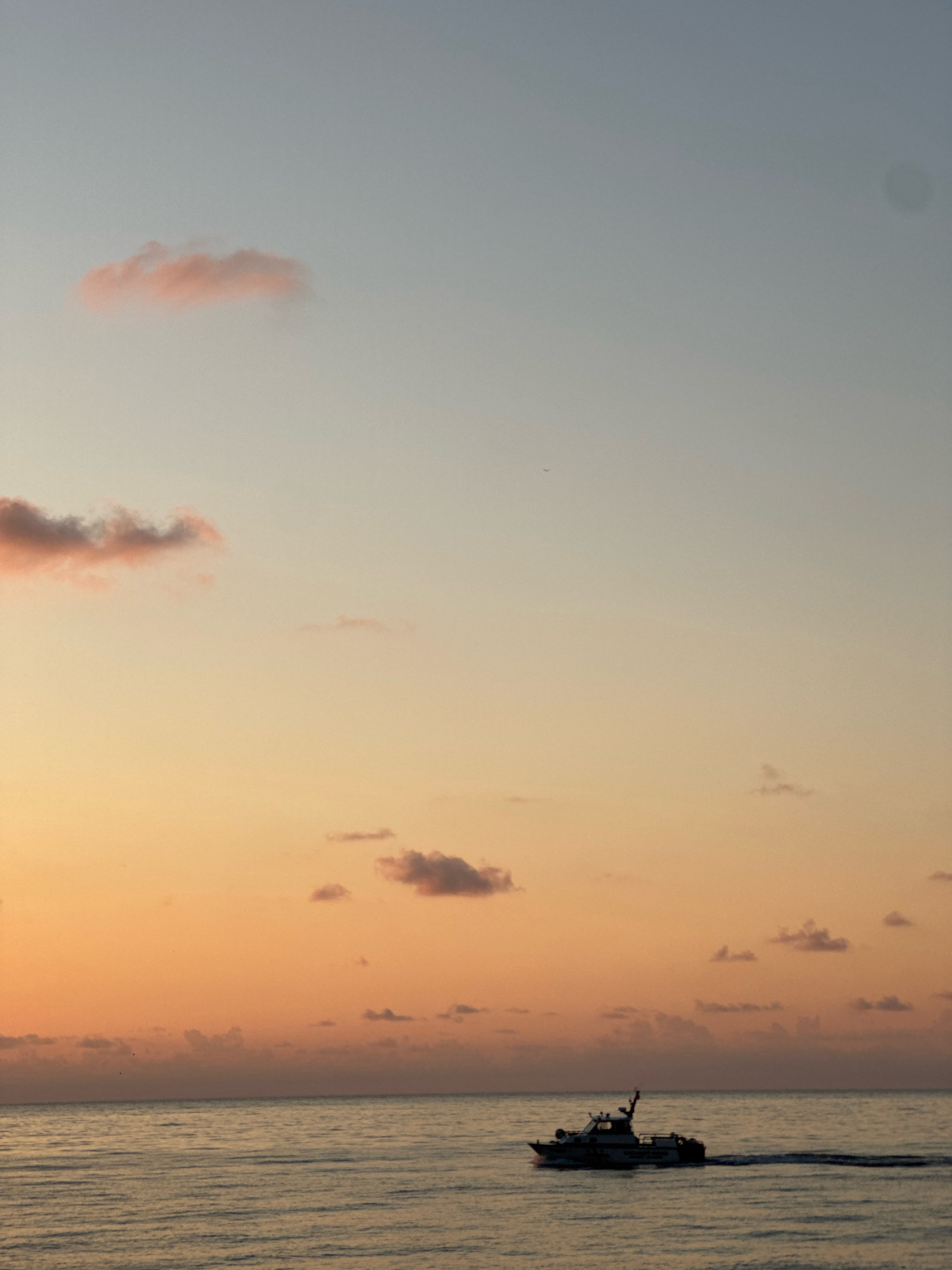 An image shows a boat moving through the water with a sunset in the background. The sky is a gradient of orange, yellow, and blue with scattered pink clouds. The water is calm and reflects the colors of the sky. The boat is white and appears to be a speed boat or yacht. It is moving from left to right in the frame, leaving a wake behind it. The overall scene is peaceful and serene. It's difficult to determine the exact location within Batumi, Georgia, without more context, but the scene evokes a coastal setting common to the city.