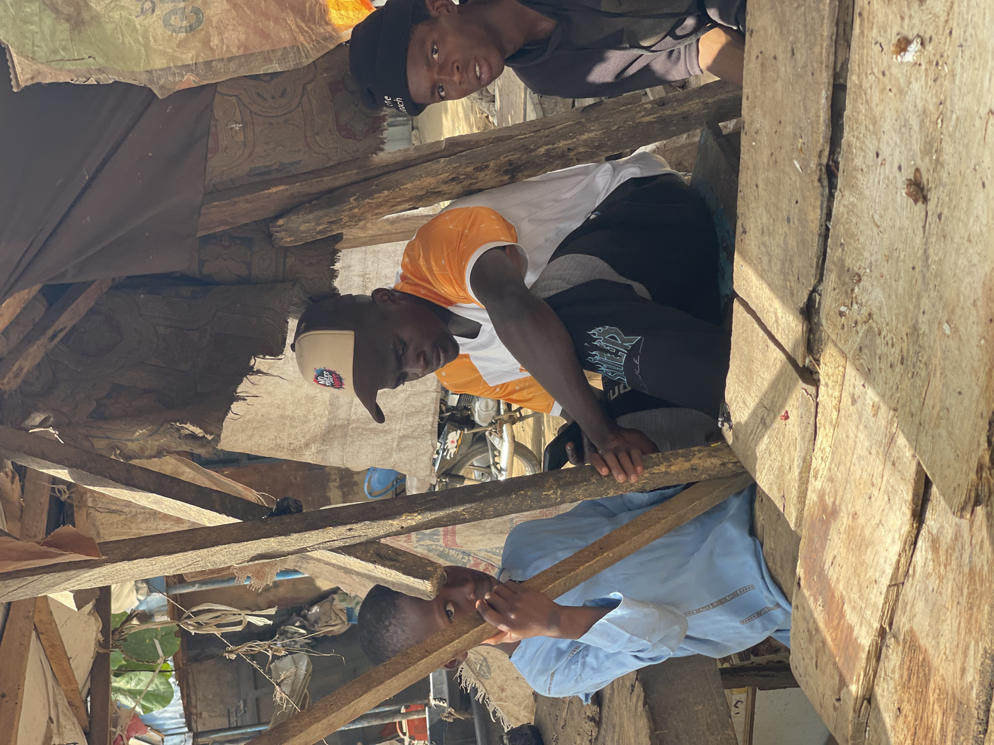 In Chikaji, Nigeria, three individuals are observed amidst a makeshift structure. One person in a light blue shirt supports a wooden beam while looking directly towards the viewer. Another person wearing a white shirt with orange sleeves works above, appearing to secure elements of the structure. A third person wearing a black beanie looks downward. The structure is made of rough wooden planks and fragments of fabric, with a glimpse of a motorcycle visible in the background.