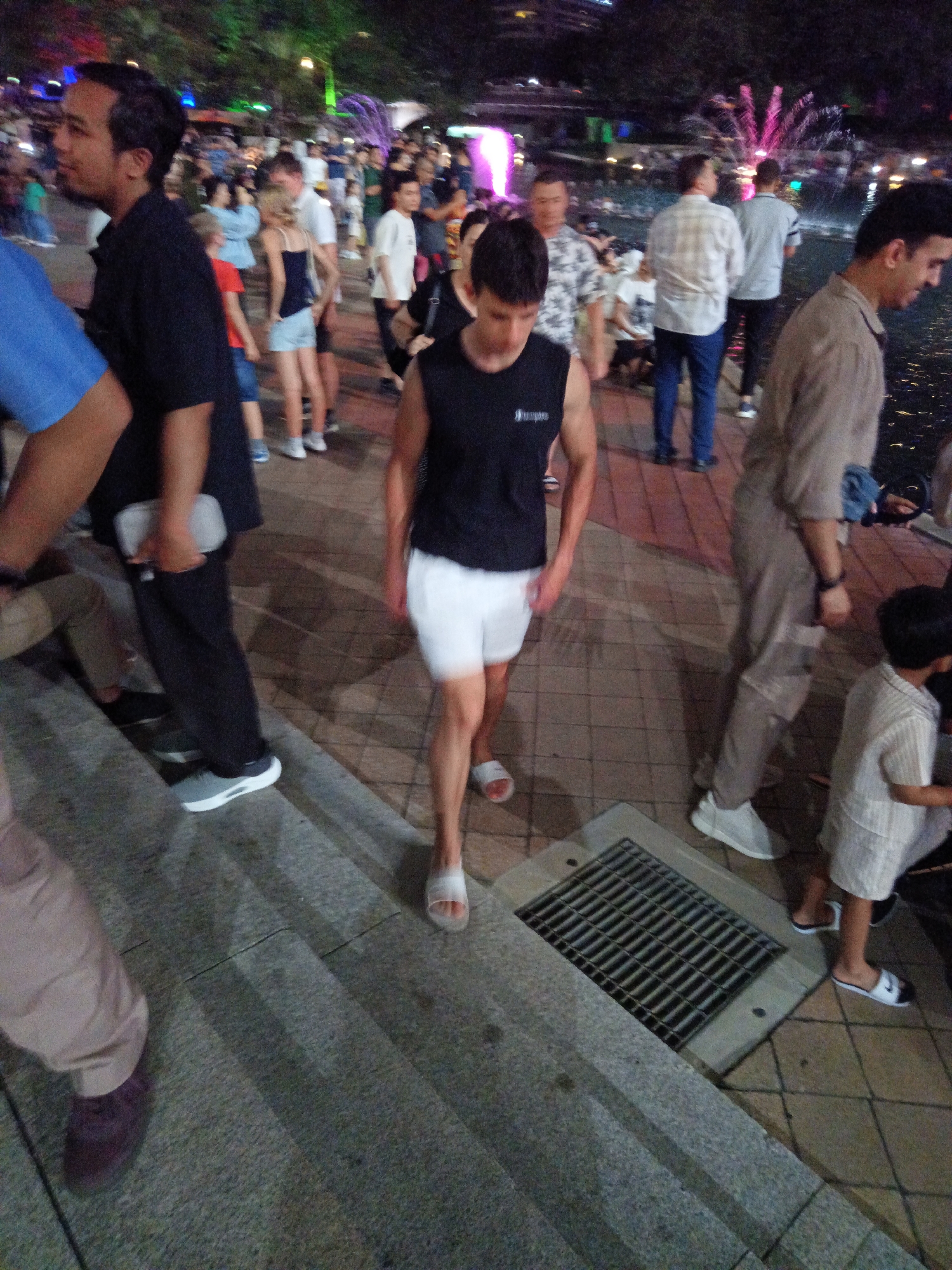 A nighttime scene depicts a public area with various people in the foreground and background. In the immediate foreground, on the left, a person wearing light brown pants and dark purple shoes is partially visible. To their right, a person in a black tank top and white shorts walks towards the viewer, with their left foot on a step. Further to the right, a person in a light brown shirt and pants walks away from the viewer. In the middle ground, a group of people are gathered, some standing and some walking. A young child in a striped shirt is visible on the far right. The background shows a body of water with a fountain and colored lights, suggesting an urban waterfront or park area. The architecture visible in the distance indicates a city environment. The overall impression is a lively, public gathering space at night.