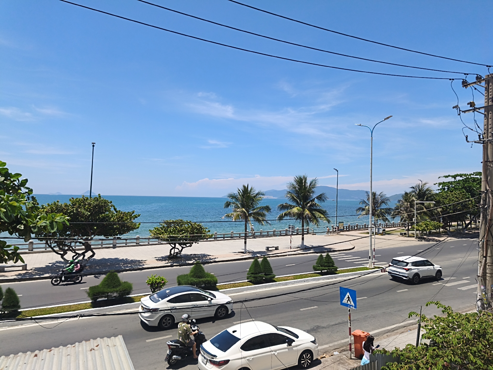 In a bright, sunlit scene with a clear blue sky, a coastal road runs alongside the ocean. Palm trees line the sidewalk, providing shade over a paved walkway and a seawall. The ocean is a vibrant turquoise, with gentle waves lapping at the shore. Beyond the ocean, hazy mountains are visible in the distance.
On the road, traffic flows in both directions. A white SUV drives on the right side of the road, crossing a pedestrian intersection. Further down the road, a white sedan is parked. In the foreground, a motorcycle with two riders, one in a patterned shirt and the other in a military-green jacket, travels along the asphalt. Another white sedan is visible behind it. A black scooter is parked at the side of the road.
On the sidewalk, a blue pedestrian crossing sign stands next to an orange trash can. A young woman in a white shirt and blue skirt is walking away from the trash can, holding a dark object. The area is populated with lush green trees and manicured hedges, adding to the tropical ambiance. The scene appears to be captured from an elevated vantage point, likely a building overlooking the street and coastline. This scene is in the city of Bac Nha Trang Ward, Vietnam.