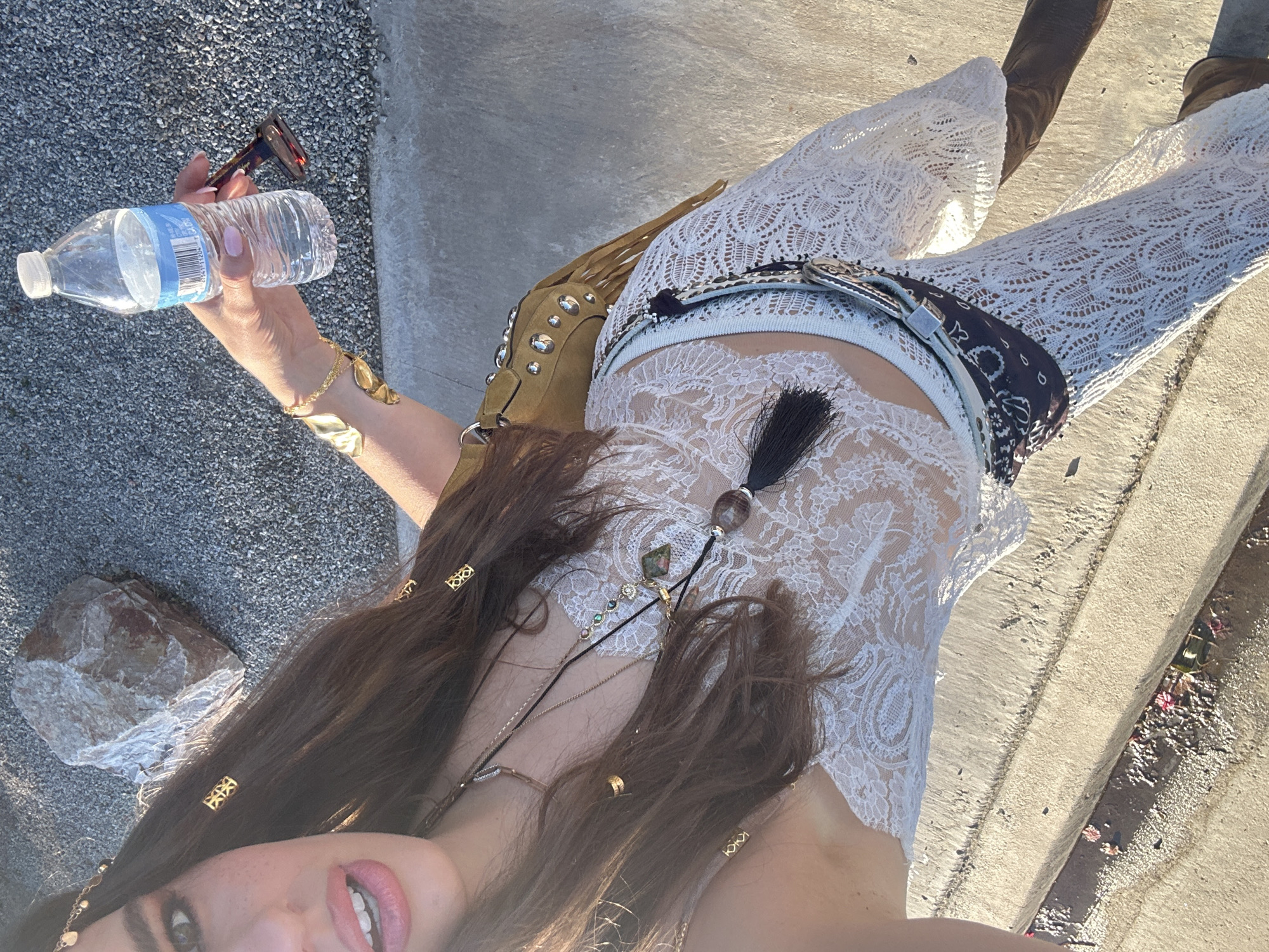 A young woman is seen from a high angle in an outdoor setting, holding a water bottle and sunglasses in her left hand. She is wearing a lacy white top and matching pants, with a brown fringed bag over her shoulder and brown boots. Her hair is long and brown, adorned with gold hair accessories and necklaces. The ground appears to be a combination of concrete and gravel. The location may be in Indio, California, United States.