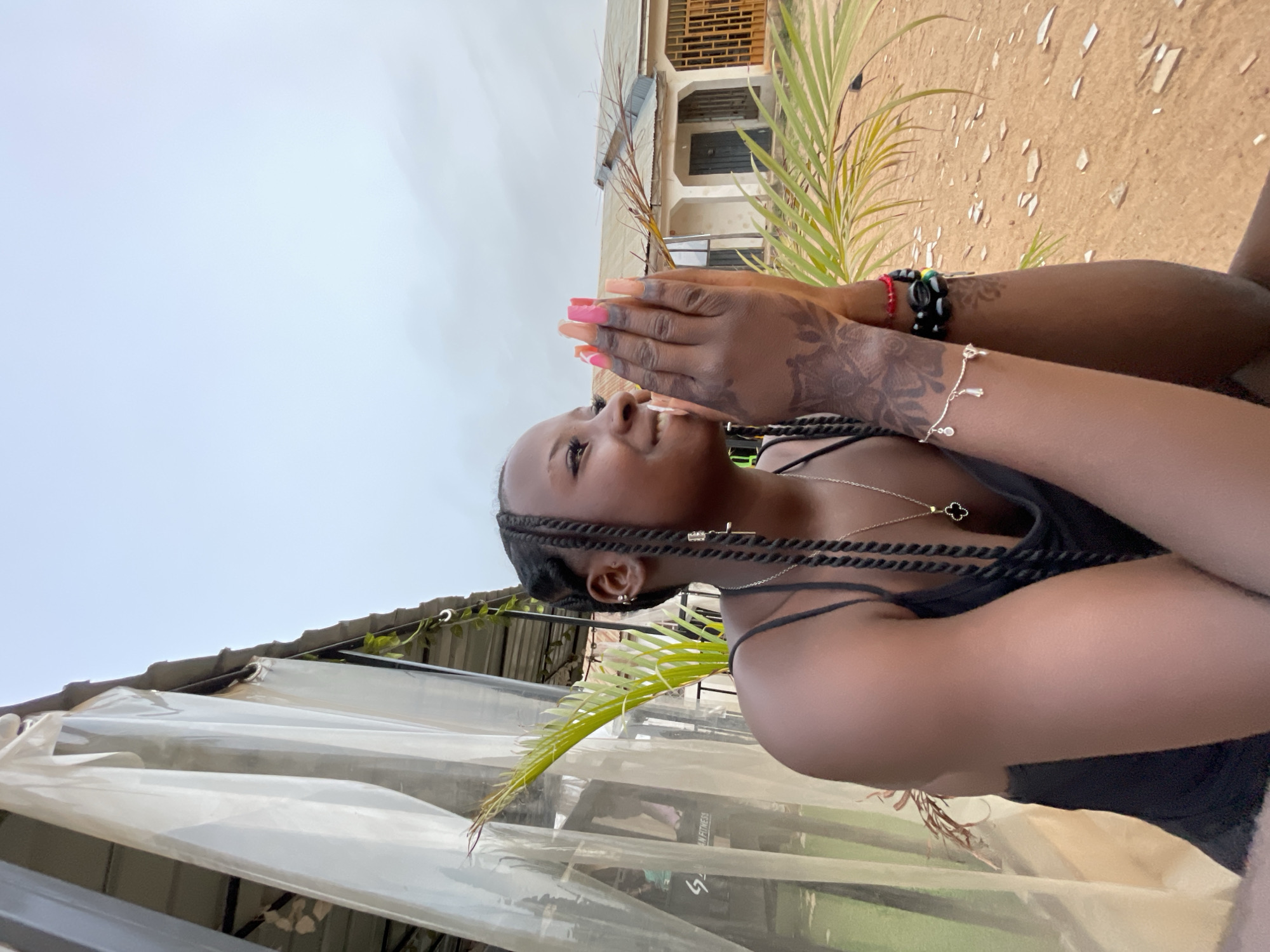 A person with dark skin, braided hair, and wearing a black tank top is holding their hands together while looking upwards. They are wearing a cross earring and a necklace with a clover pendant. The person has decorative tattoos on their hands and is wearing bracelets on one wrist. They appear to be outdoors near a building and a palm tree. Part of a translucent awning or curtain is visible in the foreground. The setting appears to be in a built-up area with sandy soil, possibly near a residential or commercial building in Jos, Nigeria.