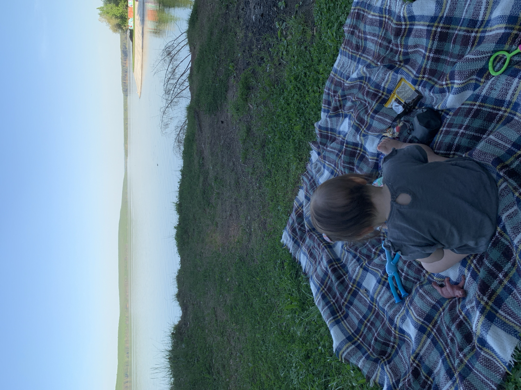 A child sits on a plaid blanket on a grassy area near the Kama River in Chaikovsky, Russia. The blanket is placed on grass next to the river bank. The child is wearing a gray shirt. Toys and other picnic items are scattered on the blanket. The river reflects the blue sky and a structure on the opposite bank. The scene appears to be a quiet outdoor setting.