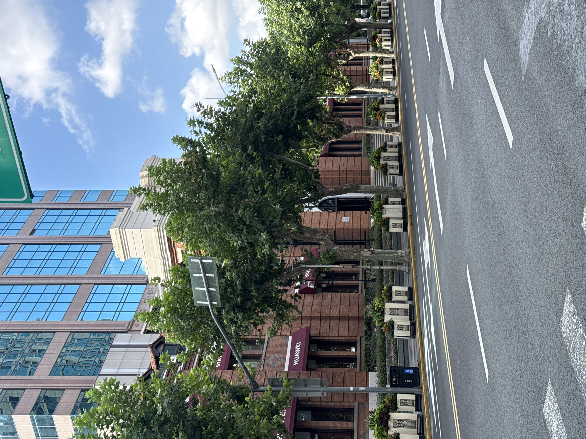 This is a street view of a city in Shanghai, China, on a bright, cloudy day. The scene captures a street with multiple lanes marked by white and yellow lines, and a pedestrian crossing. On the left side of the street, a building with a brick facade features several awnings with the "HUAWEI" logo. In front of the building, a row of trees are planted in decorative planters, with some greenery and flowers visible. Behind the brick building, a modern skyscraper with blue glass windows rises into the sky. The overall atmosphere is that of a bustling urban environment, with an emphasis on commerce and modern architecture. There are no people visible in the image.