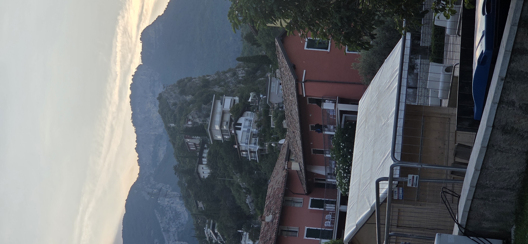 The image captures a view of Torbole, Italy, showcasing a hillside dotted with houses and a backdrop of imposing mountains. In the foreground, a white tent is set up, enclosed by metal fencing. Adjacent to the tent, a stone wall runs horizontally across the image. Beyond the tent and wall, the scene unfolds with a cluster of residential buildings, characterized by their red-tiled roofs and multi-story structures. Some of these buildings are situated on the lower slopes of the hill, while others are perched higher up, nestled amongst dense greenery. The houses vary in architectural style, with some featuring balconies and large windows. The mountainous terrain dominates the background, its rugged slopes partially covered in trees and rock formations. The sky above is a muted gray, with faint clouds hinting at either dawn or dusk. The overall impression is one of a picturesque Italian landscape, combining residential architecture with natural beauty.