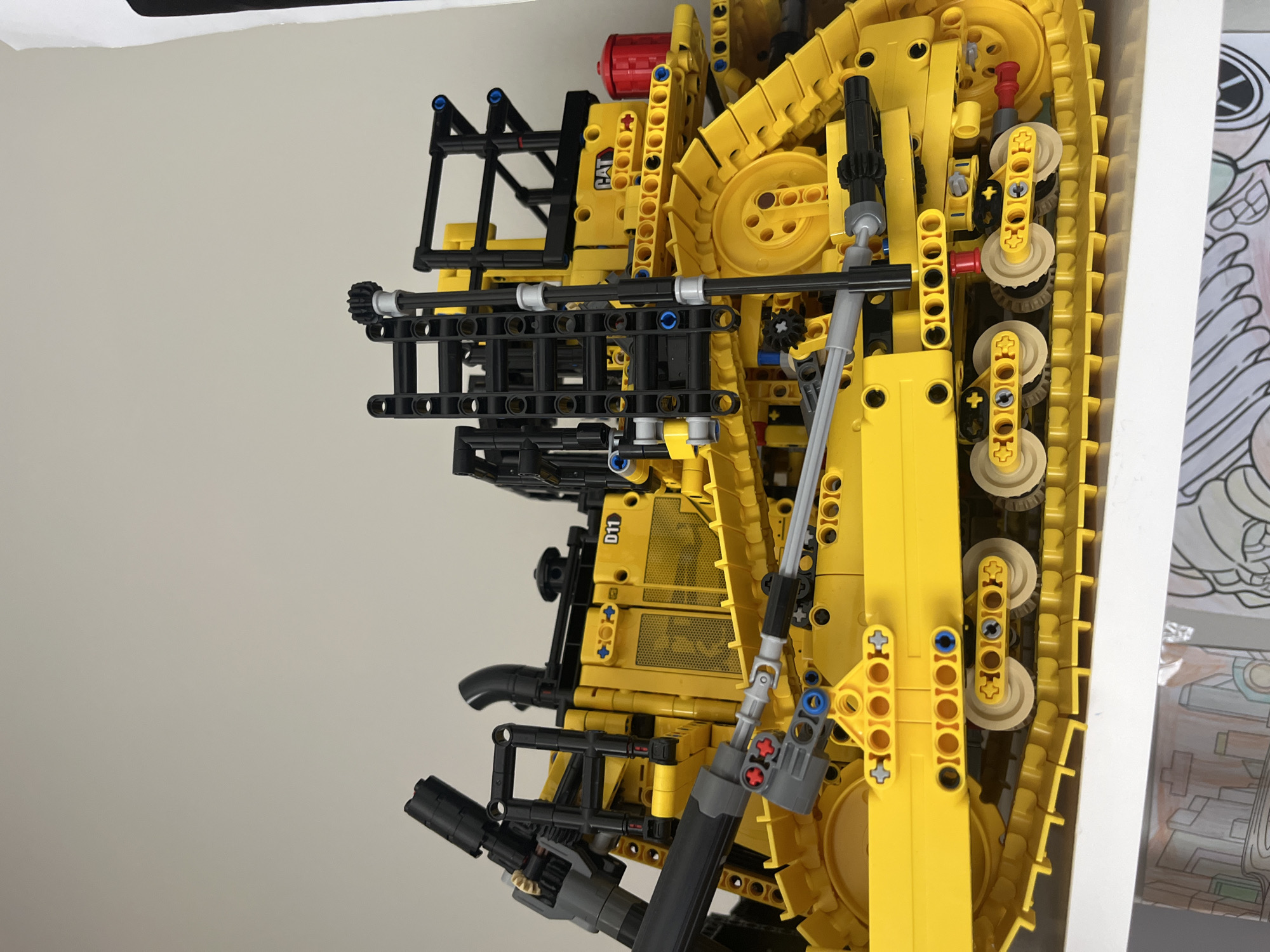 A yellow Lego Technic bulldozer, model D11 CAT, is displayed on a white shelf. The bulldozer is intricate, featuring detailed tracks, gears, and structural elements in black and gray. A red cylindrical object is visible on the right side of the bulldozer. The background is a plain, light-colored wall. The shelf is positioned above a lower surface that shows parts of colorful drawings or artwork. The image is a close-up shot, focusing on the toy. The location is in Onokivtsi, Ukraine.
