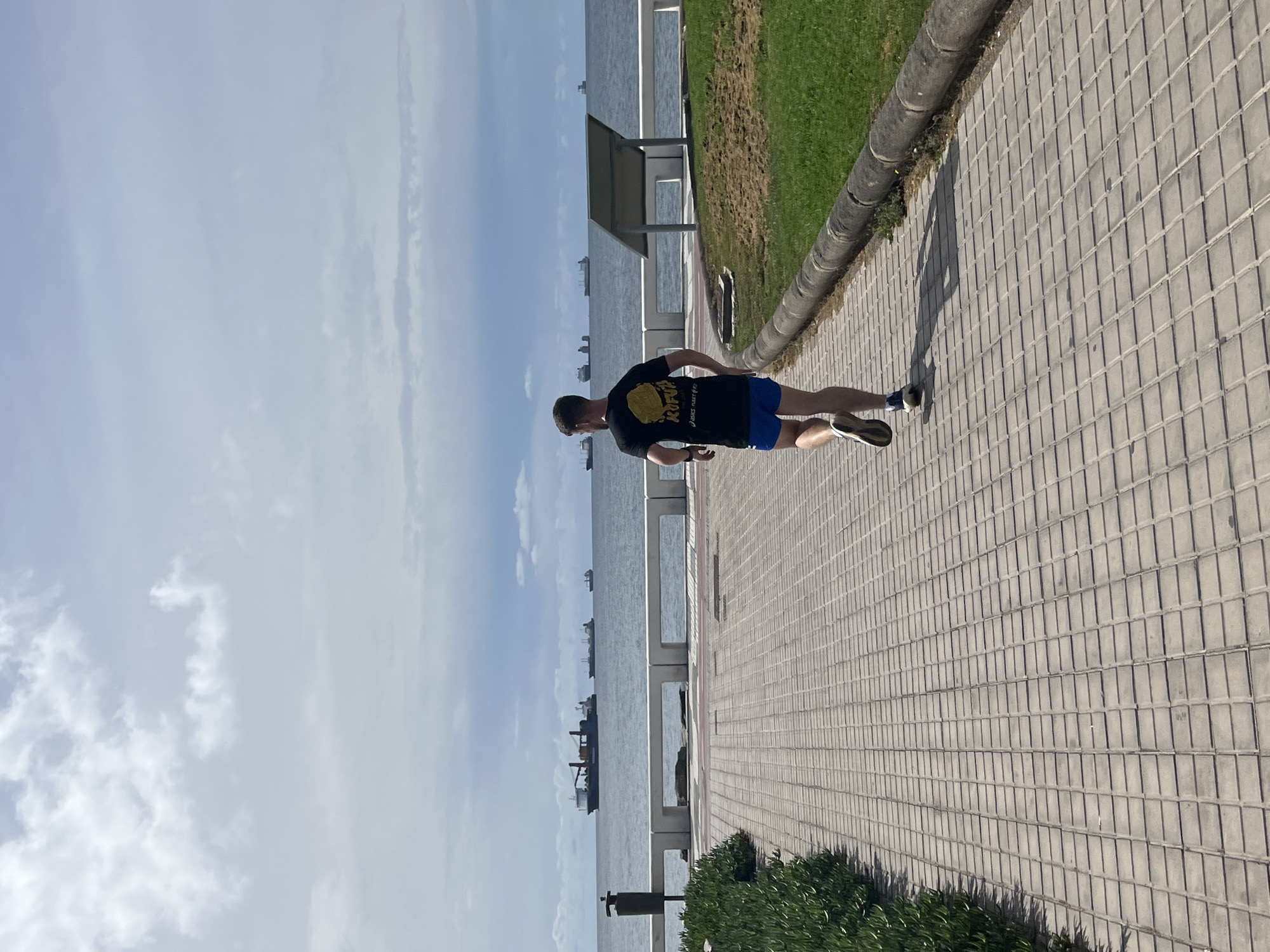 A man in a black t-shirt and blue shorts runs along a tiled sidewalk. He is next to a grassy area bordering the ocean. In the background, several ships are visible on the water. The scene is in Las Palmas, Spain, characterized by the coastal promenade.