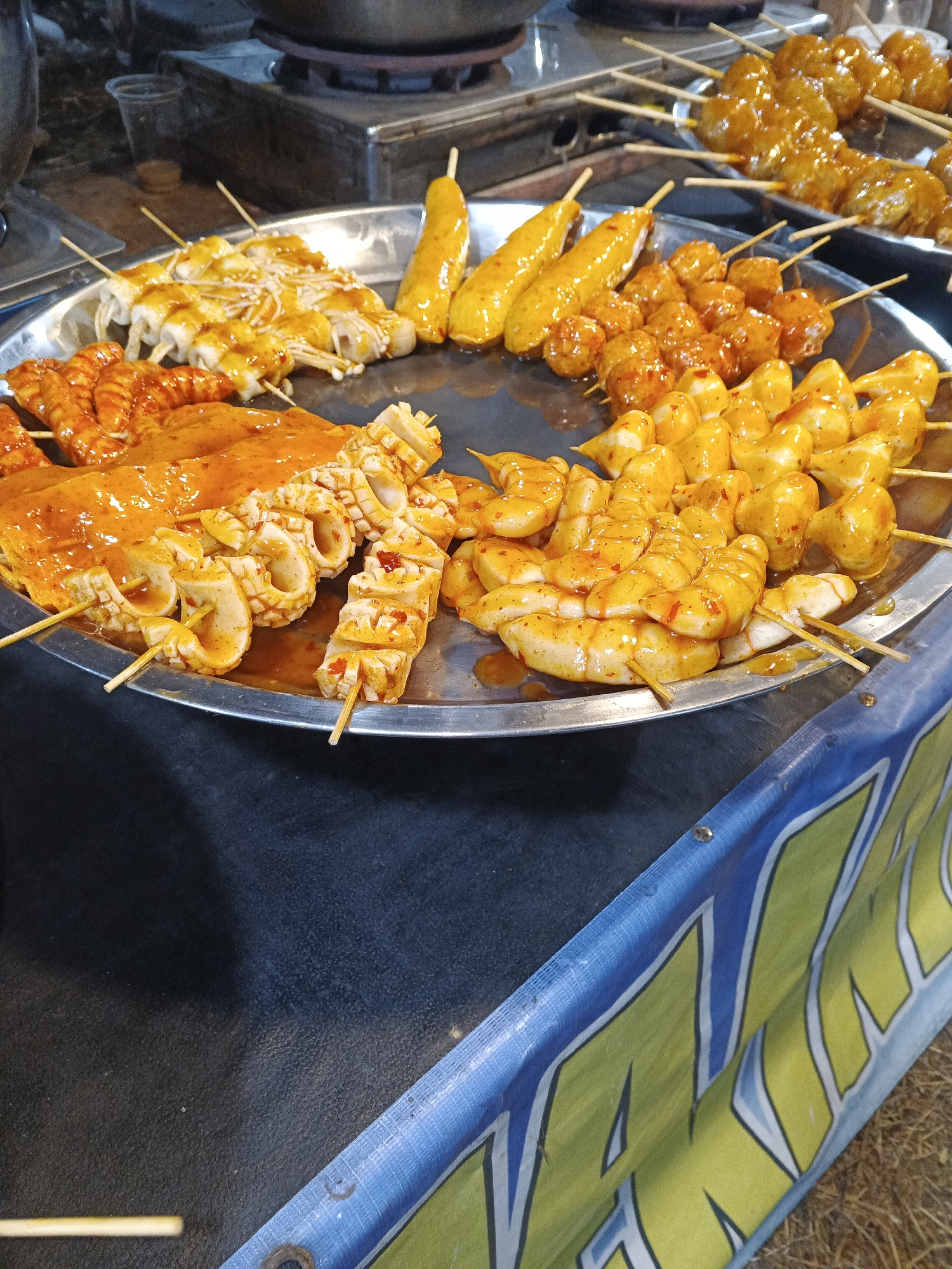 The image shows a street food stall in Sukabumi, Indonesia, featuring an assortment of skewered seafood and meat products. The food is displayed on metal platters, glistening with a bright orange sauce. Among the items are what appear to be shrimp, squid rings, and meatballs, all seasoned and ready for grilling or serving. In the background, cooking equipment and a small plastic cup can be seen, suggesting an active food preparation environment. The stall is set up outdoors, with a dark surface in the foreground and a blue and yellow banner partially visible on the right, indicating the presence of a market or street fair. The lighting suggests it might be evening or under artificial light.