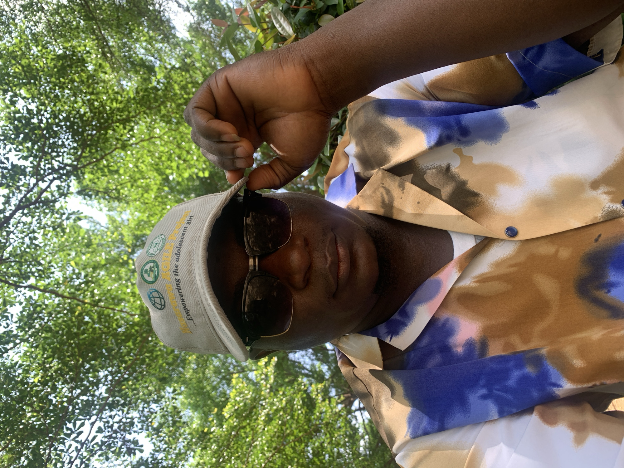 In Akwanga, Nigeria, a man is captured in a medium shot, looking directly at the camera. He is wearing sunglasses and a light-colored baseball cap adorned with circular emblems and the text "...Empowering the adolescent girl". His left hand is raised, adjusting the brim of his cap. The man is also wearing a short-sleeved shirt with a colorful tie-dye pattern featuring brown, blue, and white. The background consists of lush green foliage, suggesting an outdoor setting. The lighting indicates it is daytime.