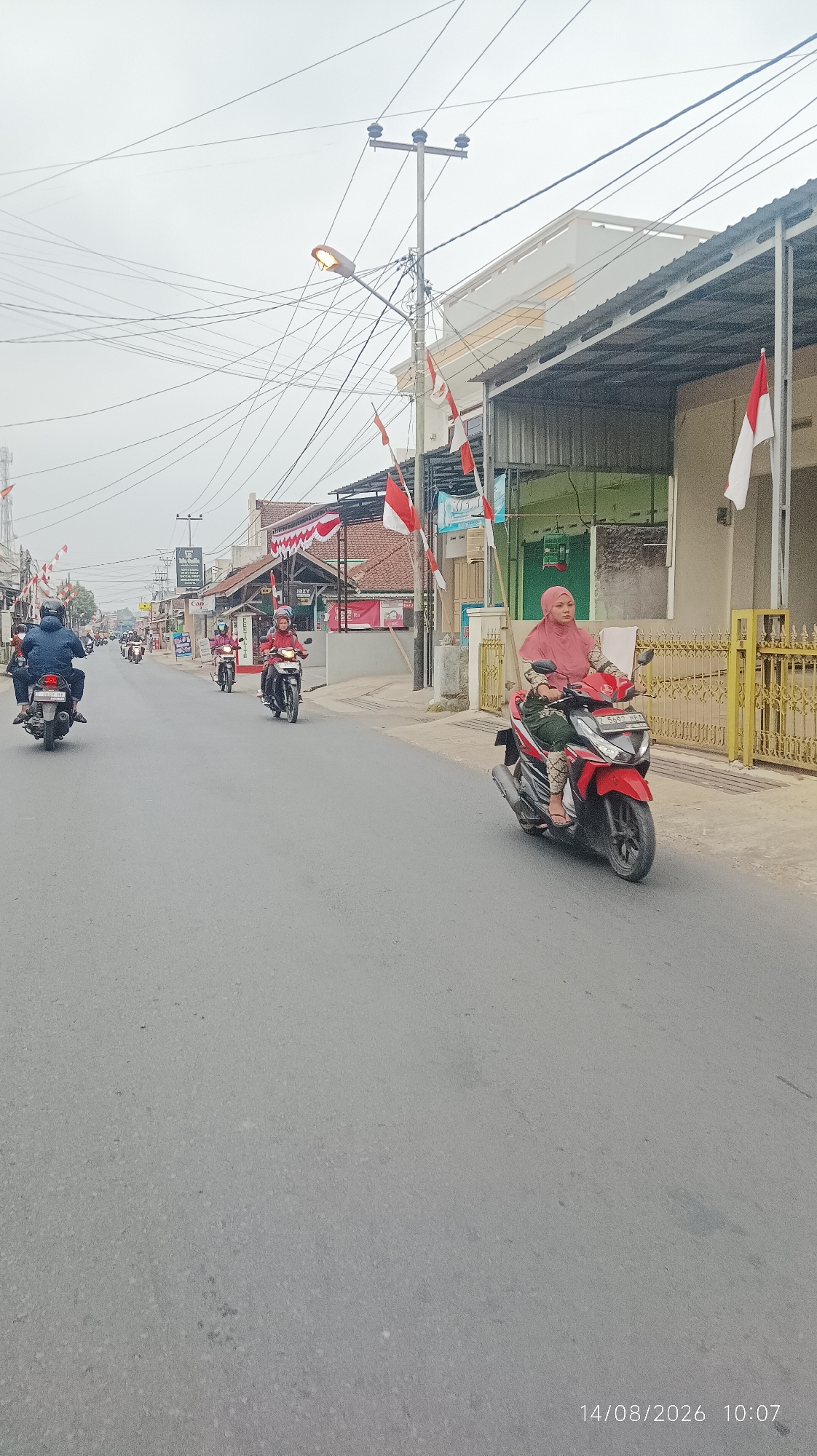 The image depicts a street scene in Tasikmalaya, Indonesia, likely during a celebration, indicated by the numerous Indonesian flags displayed. The road is paved and appears to be a two-lane street. Several people are riding motorcycles, moving in the same direction.
In the foreground, a woman wearing a pink hijab and a patterned outfit is riding a red motorcycle. To her left, another person on a motorcycle is partially visible. Further down the street, several more motorcycles are in motion, with riders wearing helmets. The buildings lining the street are of varying designs, with some featuring corrugated metal roofs and awnings. Red and white Indonesian flags are prominently displayed, strung across poles and attached to buildings, suggesting a festive or patriotic occasion. Power lines crisscross the sky, and a street lamp is visible. The sky is overcast. A timestamp in the lower right corner reads "14/08/2026 10:07".