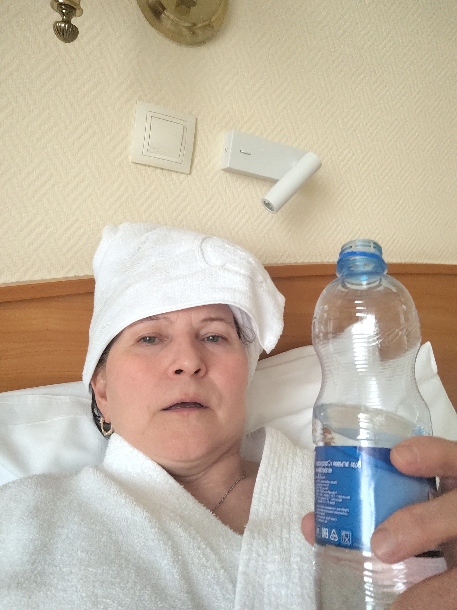 A middle-aged woman is depicted in a close-up, high-angle shot, lying in what appears to be a bed, likely in a hotel room in Moscow, Russia. She is wearing a white terry cloth bathrobe and has a matching white towel wrapped around her head like a turban. Her gaze is directed towards the camera, with a somewhat weary or neutral expression and her mouth slightly ajar. A small golden hoop earring is visible on her left ear, and a thin silver chain rests on her neck.
She holds a partially empty plastic water bottle with a blue label and cap in her right hand, which is partially visible. The text on the bottle's label is in Cyrillic script, mostly upside down and partially obscured, with phrases such as "вода питьевая" (drinking water) visible.
The background consists of a light beige wall with a subtle herringbone pattern. Mounted on the wall are a square white light switch or control panel, a modern white articulating wall-mounted spotlight, and part of a decorative metallic light fixture in the upper left corner. A wooden headboard is visible behind her. The scene suggests a moment of rest, relaxation, or possible recovery, perhaps after a shower or due to feeling unwell, with a focus on hydration.