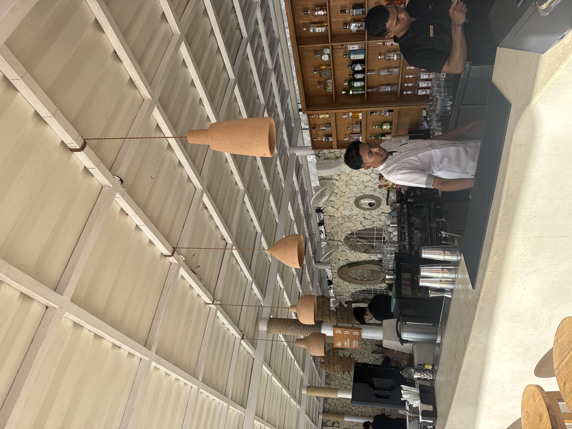 This is an interior shot of a bar in Kuta Selatan, Indonesia. The ceiling is white, with a grid-like structure from which several tan, cone-shaped lamps hang suspended by thin, brown wires.
Two men are behind the bar, one in a white shirt and the other in a black shirt. The man in the white shirt is facing forward, while the man in the black shirt is facing towards the bar, possibly preparing drinks.
Behind the bar is a wooden shelf stocked with bottles, likely alcoholic beverages. The back wall behind the shelves appears to be stone.
Various objects are visible on the bar itself, including metal cocktail shakers and other bar equipment. There are also wooden stools along the bottom-right edge of the image. The atmosphere appears bright and modern.