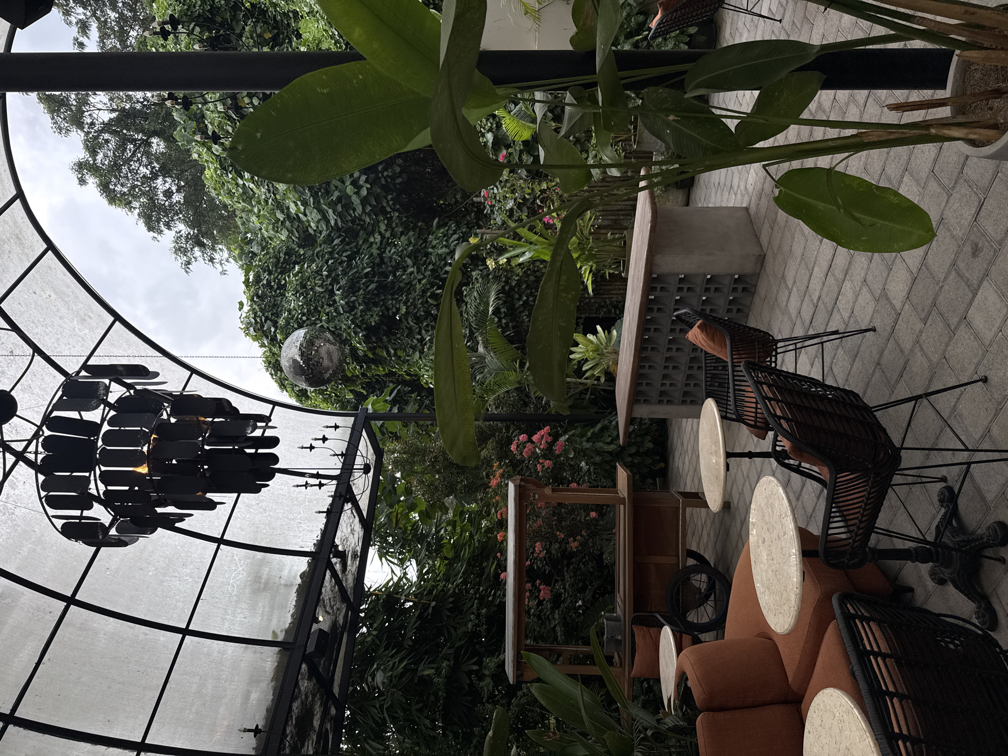 The image captures an outdoor seating area in Ubud District, Indonesia. The space is characterized by a blend of natural and constructed elements. A domed structure with a dark metal frame and translucent covering encloses a section of the seating. A large, multi-tiered chandelier hangs within this structure. Outside the dome, dense greenery including trees and various plants form a backdrop, with a disco ball suspended amidst the foliage. Seating consists of dark wicker chairs and a sofa with orange cushions, paired with small, round tables with patterned surfaces. A wooden cart and a concrete structure with decorative blocks are also visible. The ground is paved with gray, brick-like tiles. The overall scene presents a relaxed, outdoor dining or lounge area.