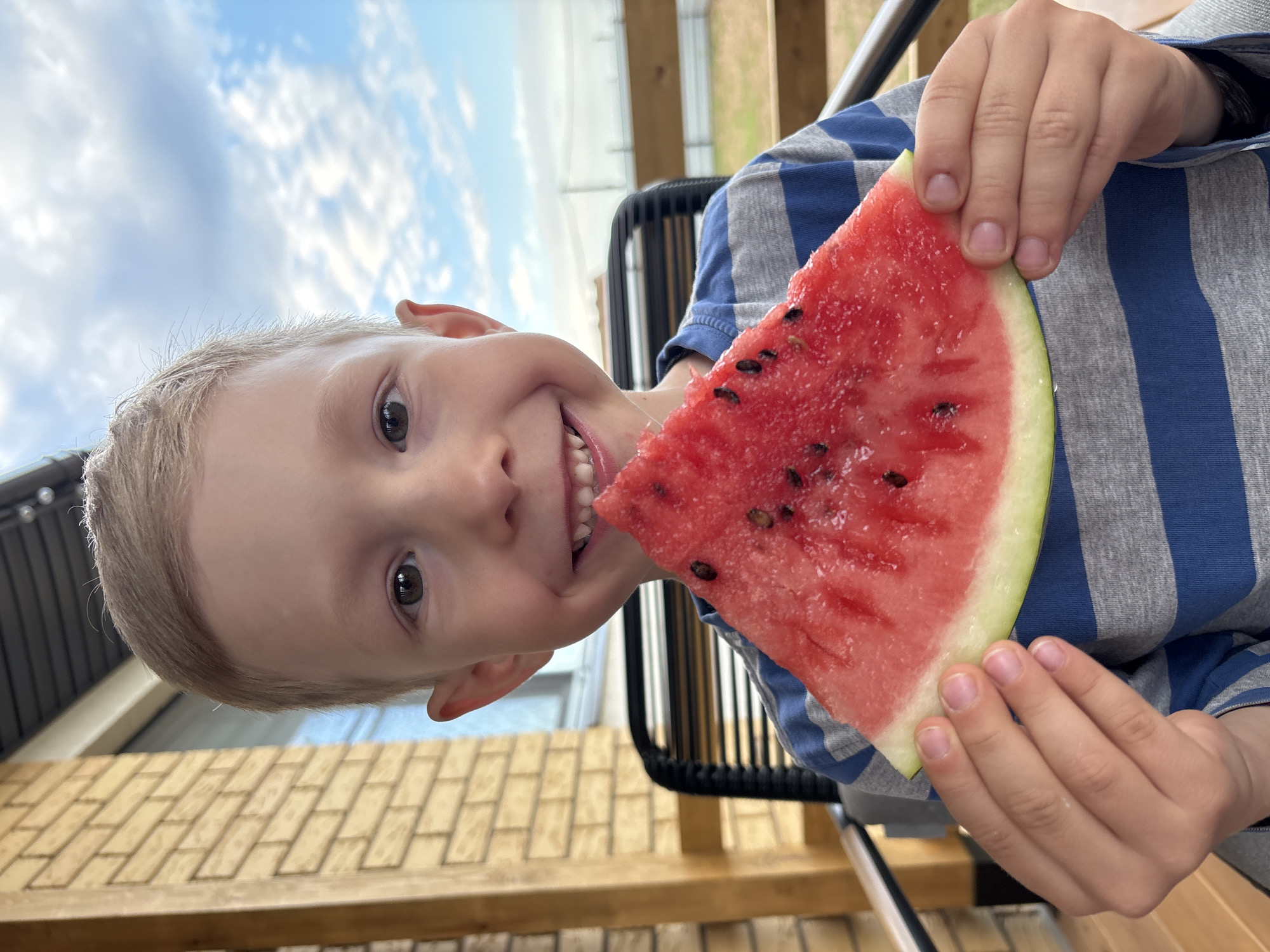 A young boy with blonde hair and fair skin is holding a slice of watermelon in front of his face. He is smiling broadly, showing his teeth, and his eyes are looking directly at the camera. He is wearing a grey and blue striped long-sleeved shirt. The background suggests an outdoor setting, possibly a porch or patio, with a brick wall and wooden structure on the left, and a glimpse of a fence and sky with clouds on the right. The image does not provide any specific information about the location in the city of Chaikovsky, Russia.