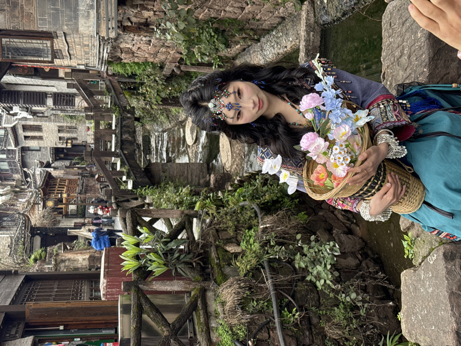 The scene depicts a young adult female seated on a stone block beside a narrow water channel in Fenghuang Ancient Town, Hunan Province, China. The woman, with long, dark, wavy hair, wears an elaborate, colorful beaded and jeweled forehead ornament with a central turquoise stone, and large blue teardrop earrings. Her eyes are accentuated with winged eyeliner. She is dressed in traditional attire, featuring a dark blue tunic with embroidered patterns in red, white, yellow, and black, adorned with multiple silver-colored bangles and beaded fringes on her sleeves. She wears a teal-colored skirt. She holds a woven bamboo basket filled with artificial flowers, including white, pink, orange, and blue varieties, looking directly at the viewer with a neutral expression.
To her left, a wooden railing with crisscrossing supports runs alongside the water channel, which contains shallow water and visible circular stone stepping pads. The banks are lined with rough-hewn stone and covered in dense green and dried foliage. In the mid-ground, a small wooden arched bridge spans the water channel. Beyond the channel and bridge, a narrow street is visible, populated by several individuals, including one person in a blue jacket. The background features multi-story traditional Chinese buildings constructed from gray stone and dark wood, characterized by intricate architectural details and dark tiled roofs, typical of Fenghuang's ancient architecture. A partial view of a hand is visible in the lower right foreground, suggesting a person is taking the photograph.