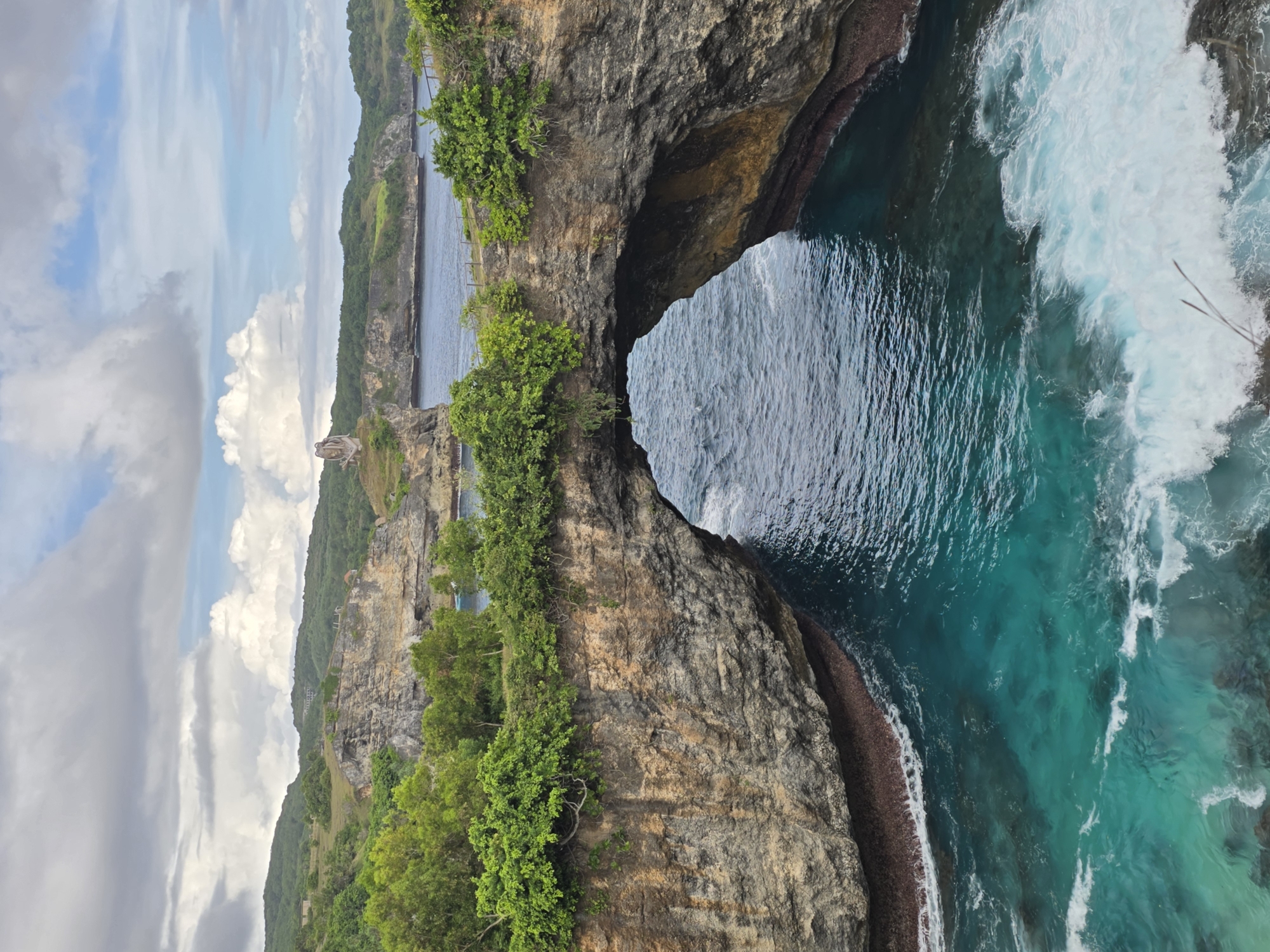 The image shows a rocky cliff formation in Nusa Penida, Indonesia, with a natural archway carved out by the sea. Green vegetation clings to the cliff's surface. The turquoise water of the ocean is visible through the arch and on the right side of the image, where waves are crashing against the rocks. In the background, a coastline can be seen, with more green vegetation and a few buildings. The sky is partly cloudy with patches of blue.