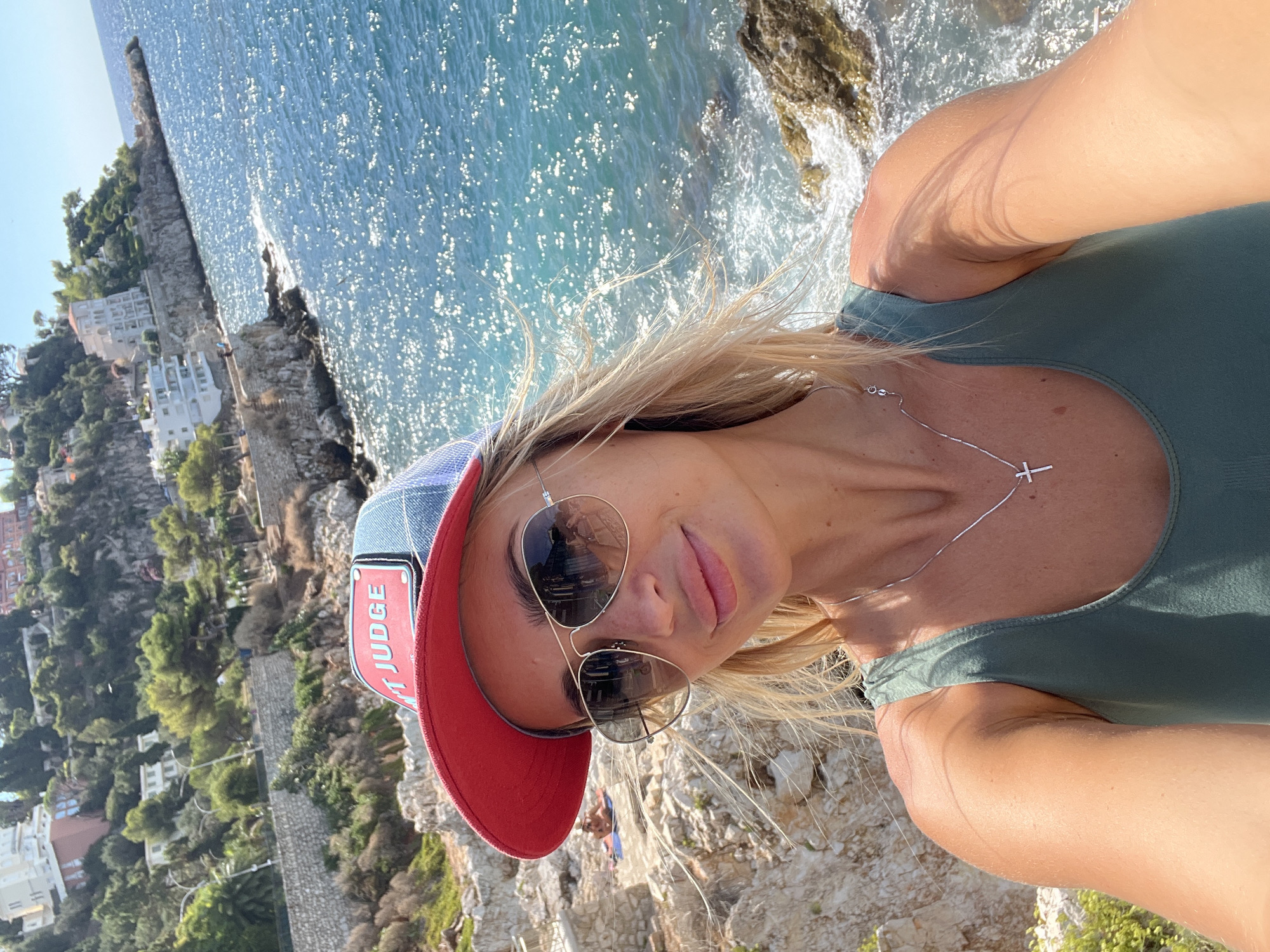 A woman with blonde hair, wearing a green swimsuit and a red and blue trucker hat, takes a selfie on a rocky coastline. Her sunglasses reflect the sunlit blue water of the Mediterranean Sea. In the background, buildings are visible on a hillside overlooking the sea. The scene is set in Nice, France, likely along the coastal path or a rocky beach area.