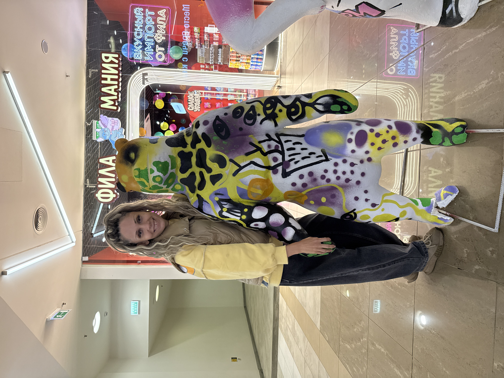In the city of Yaroslavl, Russia, a woman with long, blonde, curly hair, styled in a high ponytail, stands in a brightly lit indoor shopping area. She is smiling at the camera and is positioned to the left of a large, stylized bear sculpture. The sculpture is painted in vibrant colors and features abstract patterns and graffiti. The woman is wearing a yellow long-sleeved shirt, a beige vest, and dark wash denim jeans. She has on light brown athletic shoes. The background reveals a store with neon signs, one reading "Вкусный импорт от фила" (Tasty import from Phila), and another with Cyrillic text that is partially visible. The floor is tiled with a reflective surface. An exit sign is visible on the wall in the upper left corner.
