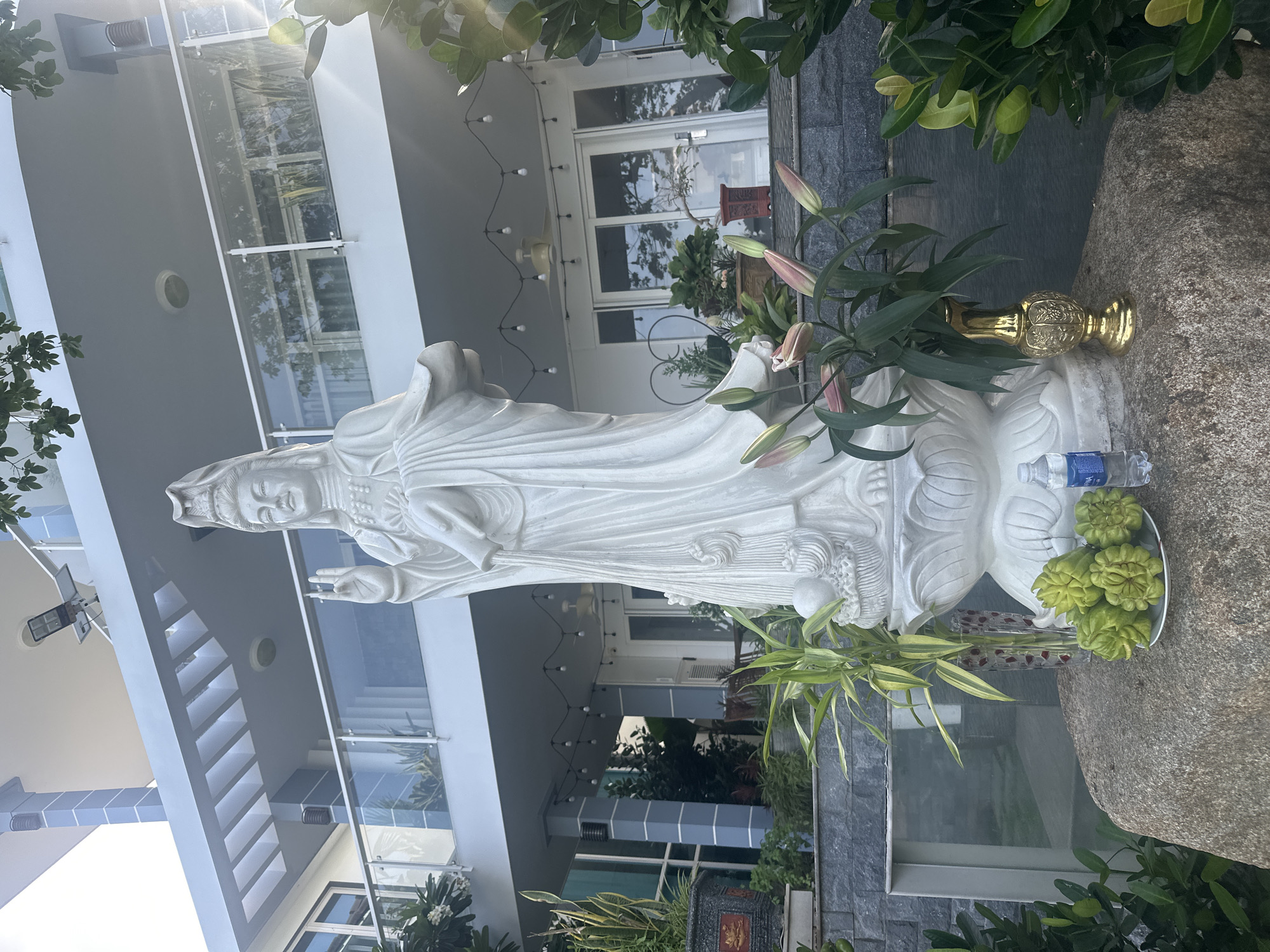 A white marble statue of a Buddhist deity, likely Guanyin, stands prominently on a large rock in front of a multi-story modern house in Nam Nha Trang Ward, Vietnam. The statue, with its flowing robes and serene expression, is the focal point. At the base of the statue, on the rock, are offerings including a plate of green, bulbous fruits, a golden vase with flowers, and a bottle of water. The house behind the statue features a contemporary design with white walls, large glass windows, and a balcony with a glass railing. String lights are visible under the overhang of the house, and potted plants are strategically placed around the property, adding greenery to the scene. The overall impression is one of a tranquil, spiritual space within a residential setting.