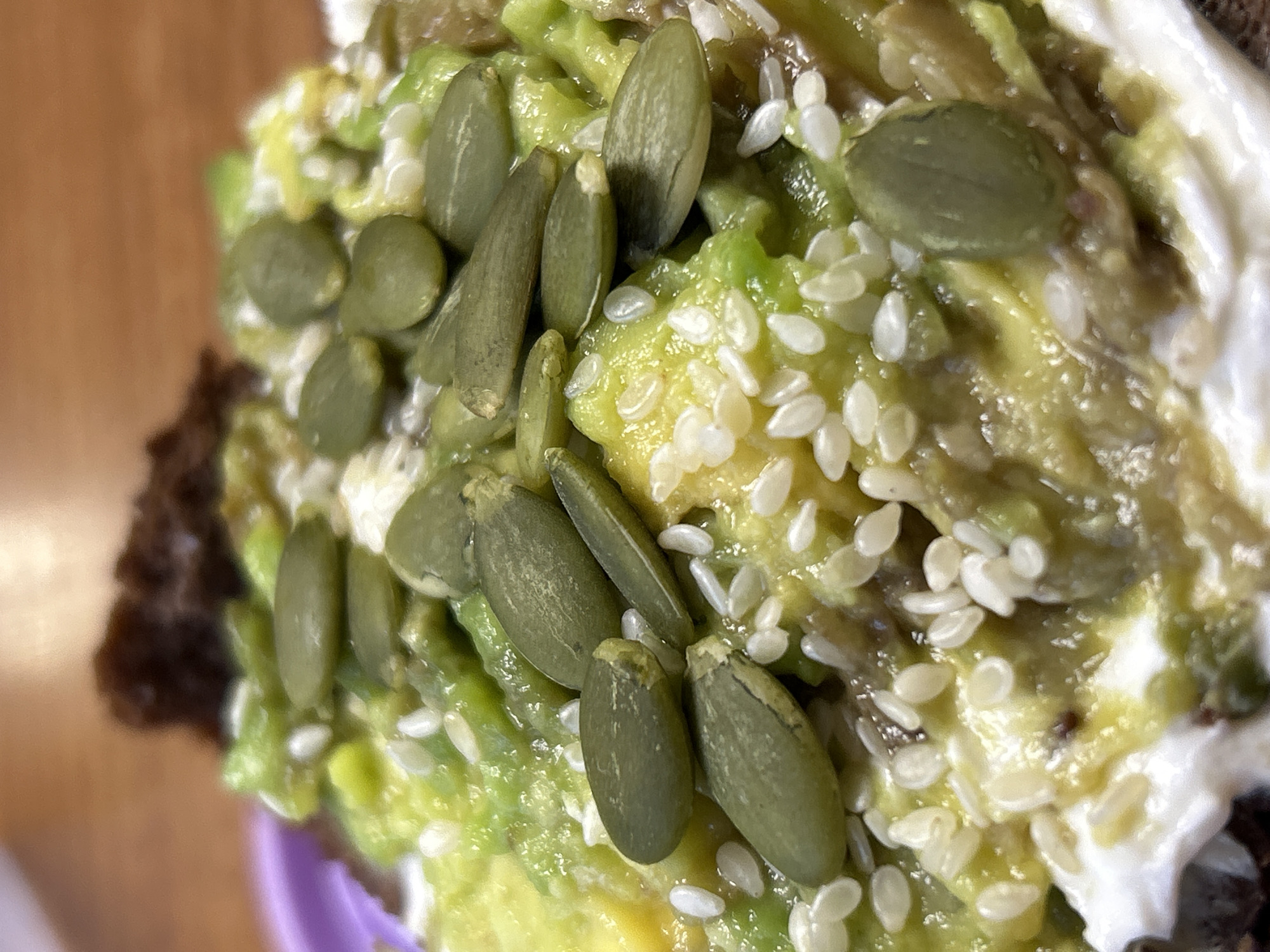 A close-up shot shows an avocado toast with a topping of pumpkin seeds and sesame seeds. It sits on what appears to be dark bread with a spread of avocado and a white creamy substance. The setting is not discernible, and there's no indication of the location being Ural'skoe, Russia from the image alone.