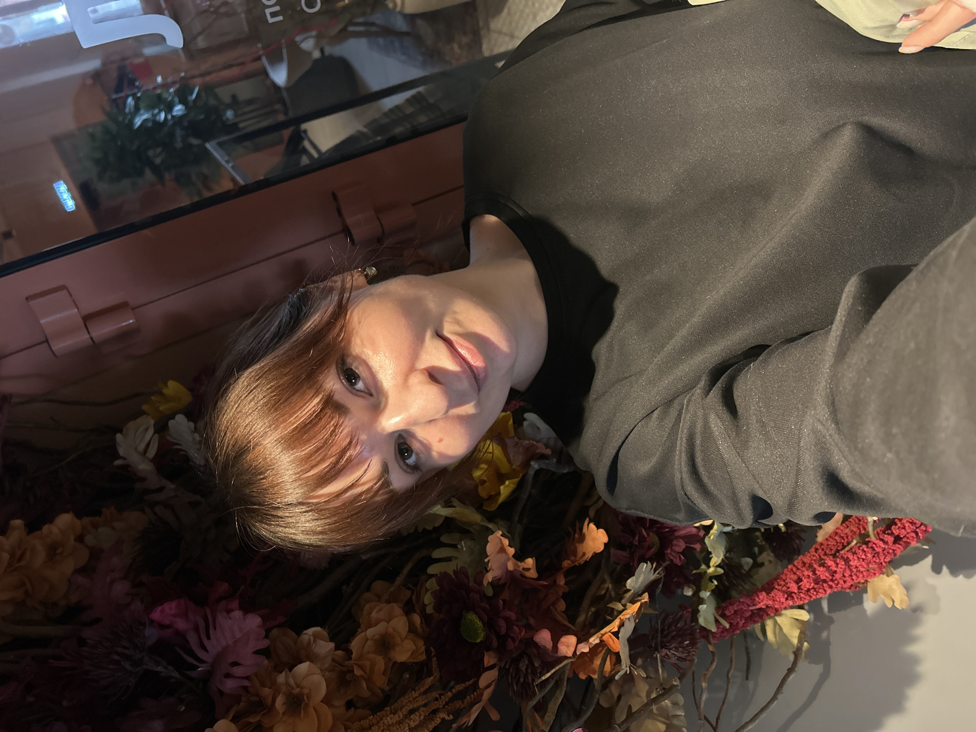 The image depicts a female subject captured in a selfie indoors. She has light-brown hair with bangs, fair skin, and is looking directly at the camera with a slight smile. She is wearing a dark, long-sleeved top. A small, light-colored earring is visible in her right ear. Her right hand, displaying manicured nails, is partially visible in the bottom right corner, suggesting she is holding the camera.
The background to the left of the subject is dominated by a dense arrangement of artificial flowers and foliage, displaying colors such as deep reds, burgundies, oranges, yellows, and creams. A red, textured, possibly woven, decorative element is visible at the lower left of this floral display.
To the right and behind the subject, a glass door or window is visible, framed by a reddish-brown paneled wall structure. A blue, illuminated "ВЫХОД" (EXIT) sign is clearly reflected on the glass, indicating a public indoor space in Russia. Additional Cyrillic letters, including "Л," "по," and "о," are partially legible on the glass, likely part of a business name. Through the glass, a large potted plant with dark green leaves and a curved, light-colored object (possibly a piece of furniture) are discernible. The precise location within the city of Chaikovsky, Russia, cannot be determined from the visual evidence provided.