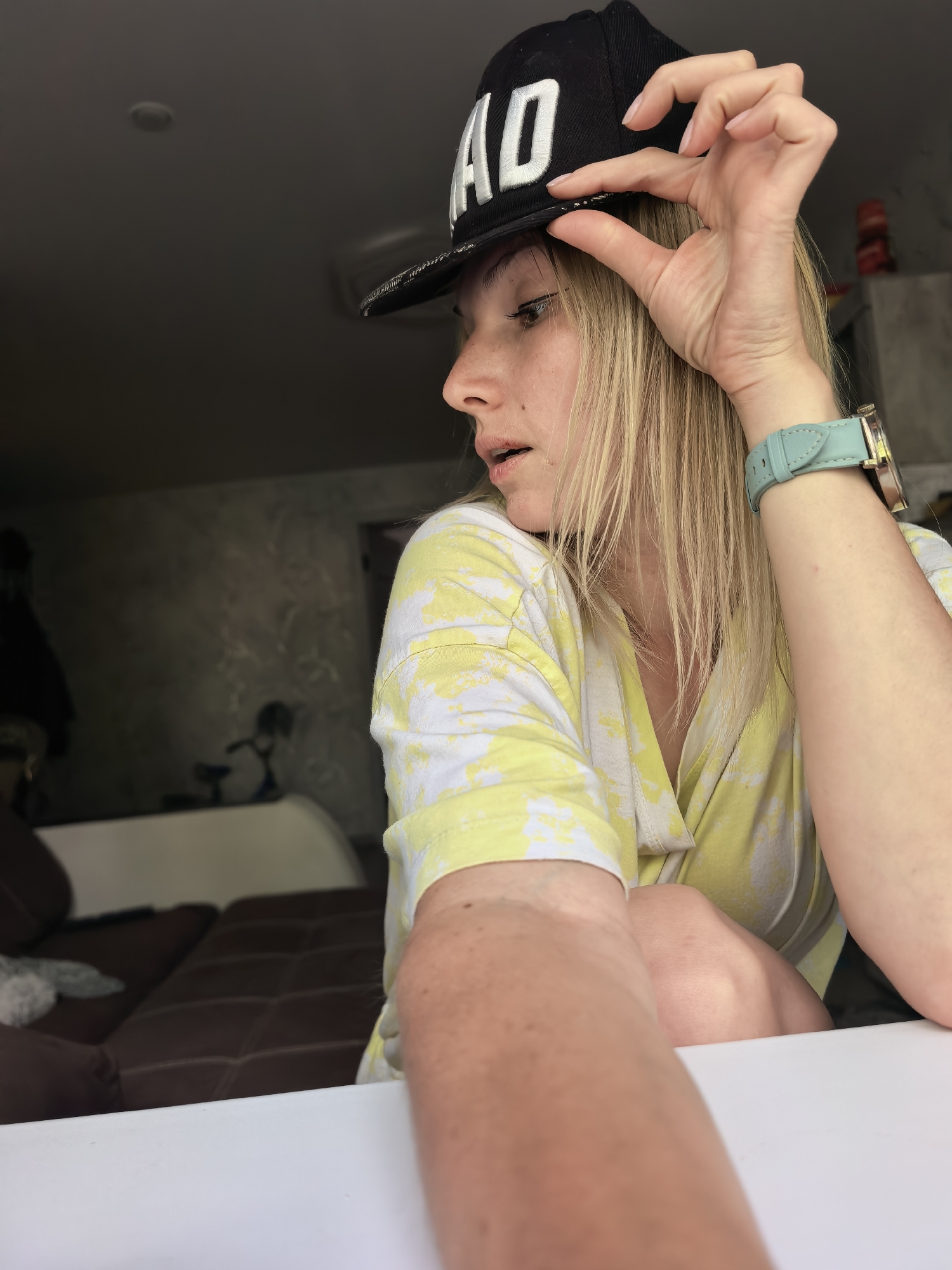 A woman is indoors, wearing a black baseball cap with "AD" in white lettering, a yellow and white tie-dye short-sleeved shirt, and a light-blue watch. She is holding the brim of her cap with her right hand. She is fair-skinned with blonde hair. A brown couch is visible in the background. A white table is in the foreground. The location is Sterlitamak, Russia.