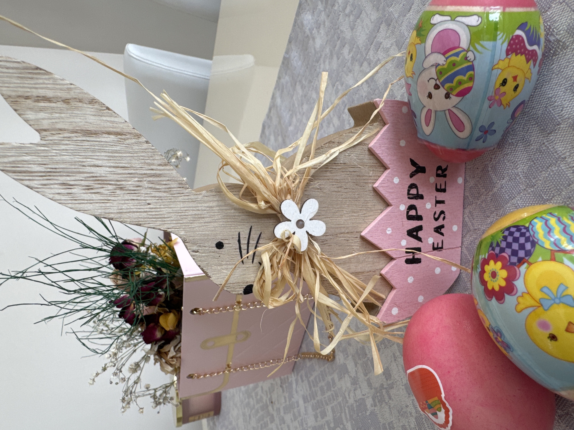 The image shows a close-up of Easter decorations arranged on a grey surface. The decorations include a wooden rabbit head with long ears, adorned with a dried flower arrangement and a raffia bow. To the right of the rabbit head, there are painted Easter eggs in various colors and patterns. A pink wooden sign with the words "Happy Easter" is also visible. The background is slightly blurred, but it appears to be a neutral-toned interior, possibly a table setting. The image does not contain sufficient information to determine its location in Varna, Bulgaria or involve any people, subjects, actions or interactions.