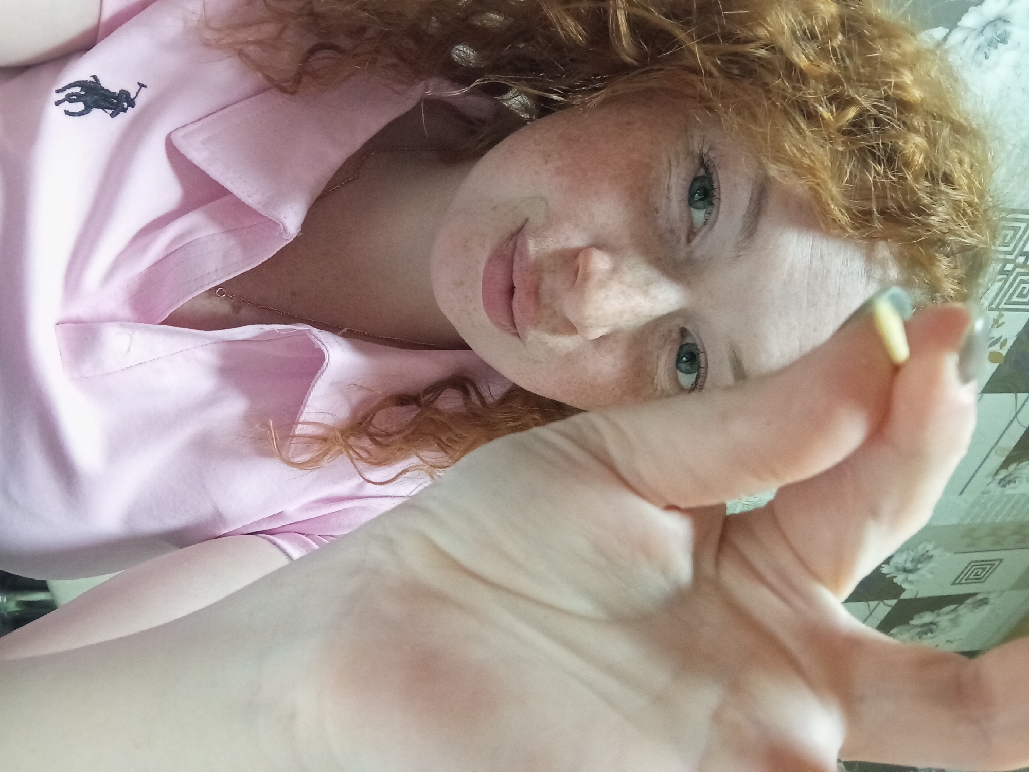 A young woman is depicted in a close-up, high-angle shot, likely lying down and looking upwards towards the camera. She has distinct red, curly hair, fair skin, and numerous freckles across her face. Her eyes are a light, striking green or blue-green, and she sports a gentle, relaxed smile.
She is wearing a light pink, collared shirt with the black Ralph Lauren polo logo (a polo player on a horse) embroidered on the left side of the chest. A thin, delicate metallic necklace is visible around her neck. Her right hand is extended prominently into the foreground, slightly out of focus, with fingers spread. Faint traces of light-colored nail polish (yellow and gray) can be seen on some of her fingernails.
The background on the right side of the frame shows a light-colored, patterned textile, possibly a bedspread or tablecloth, with a combination of geometric and floral designs. The lighting appears soft and even, typical of an indoor setting.
The woman's gaze is directed straight at the viewer, conveying a calm and friendly emotion. The only visible text is the Ralph Lauren logo on her shirt. The setting appears to be an interior space, without specific cues to indicate the time of day or weather beyond indoor conditions. The location context for this image is Yurgamysh, Russia.