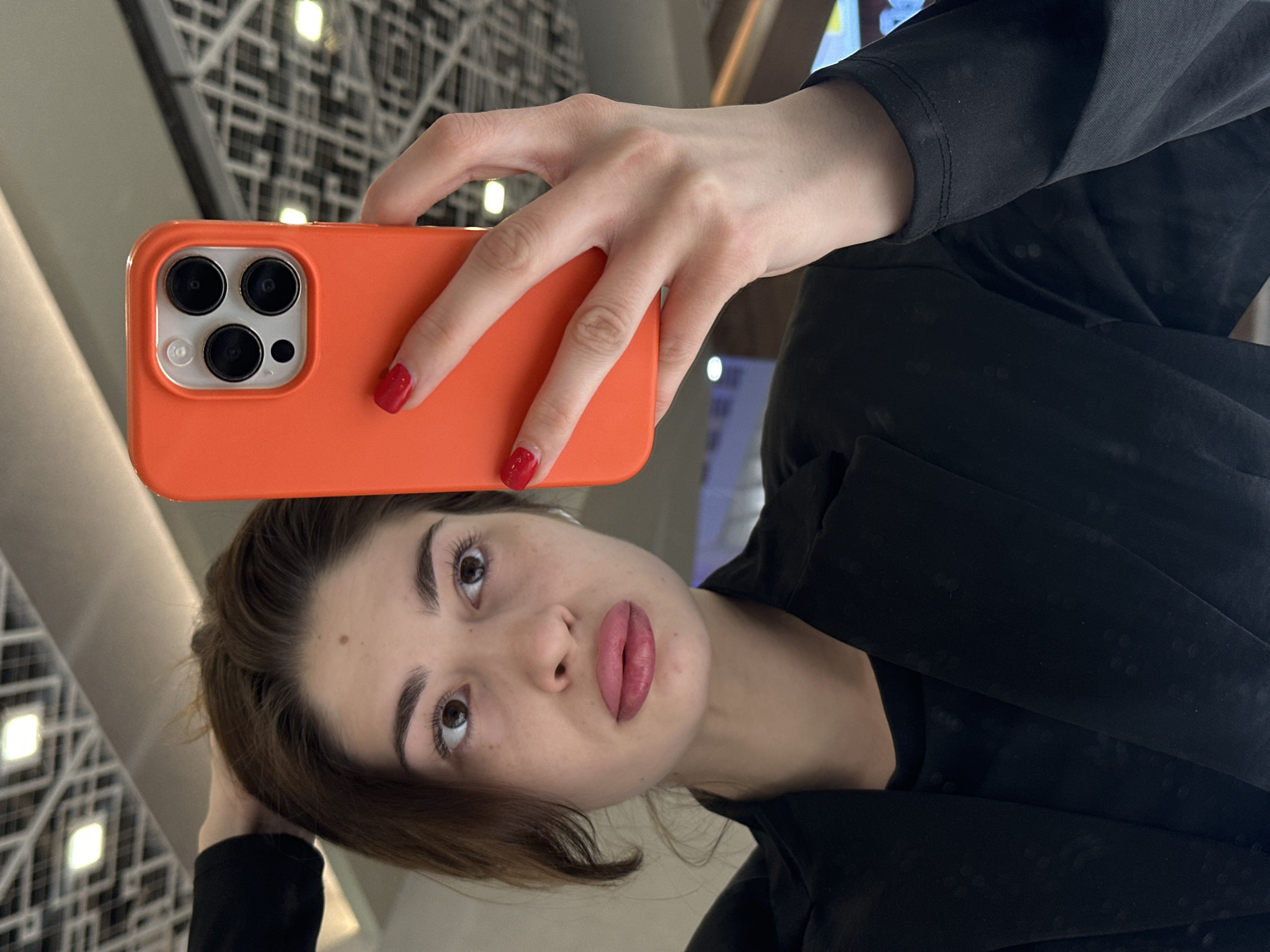 The image depicts a young adult female taking a selfie in what appears to be an indoor commercial or public facility located in Yekaterinburg, Russia. The specific location within Yekaterinburg is not identifiable from the visual information. The subject, appearing to be in her 20s or early 30s, has brown hair and brown eyes, and her lips are notably full and tinted pink. She is wearing a black long-sleeved garment underneath a black blazer or jacket. Her right hand, adorned with red nail polish, holds a silver-colored smartphone encased in a bright orange protective cover, which features a prominent triple-lens camera module. Her left hand is partially visible near the top of her head, seemingly adjusting her hair. Her gaze is directed slightly upward and to the right. The background, likely a reflection, shows a modern interior ceiling structure with a grid pattern and integrated rectangular lighting units, consistent with a shopping mall or large retail establishment.