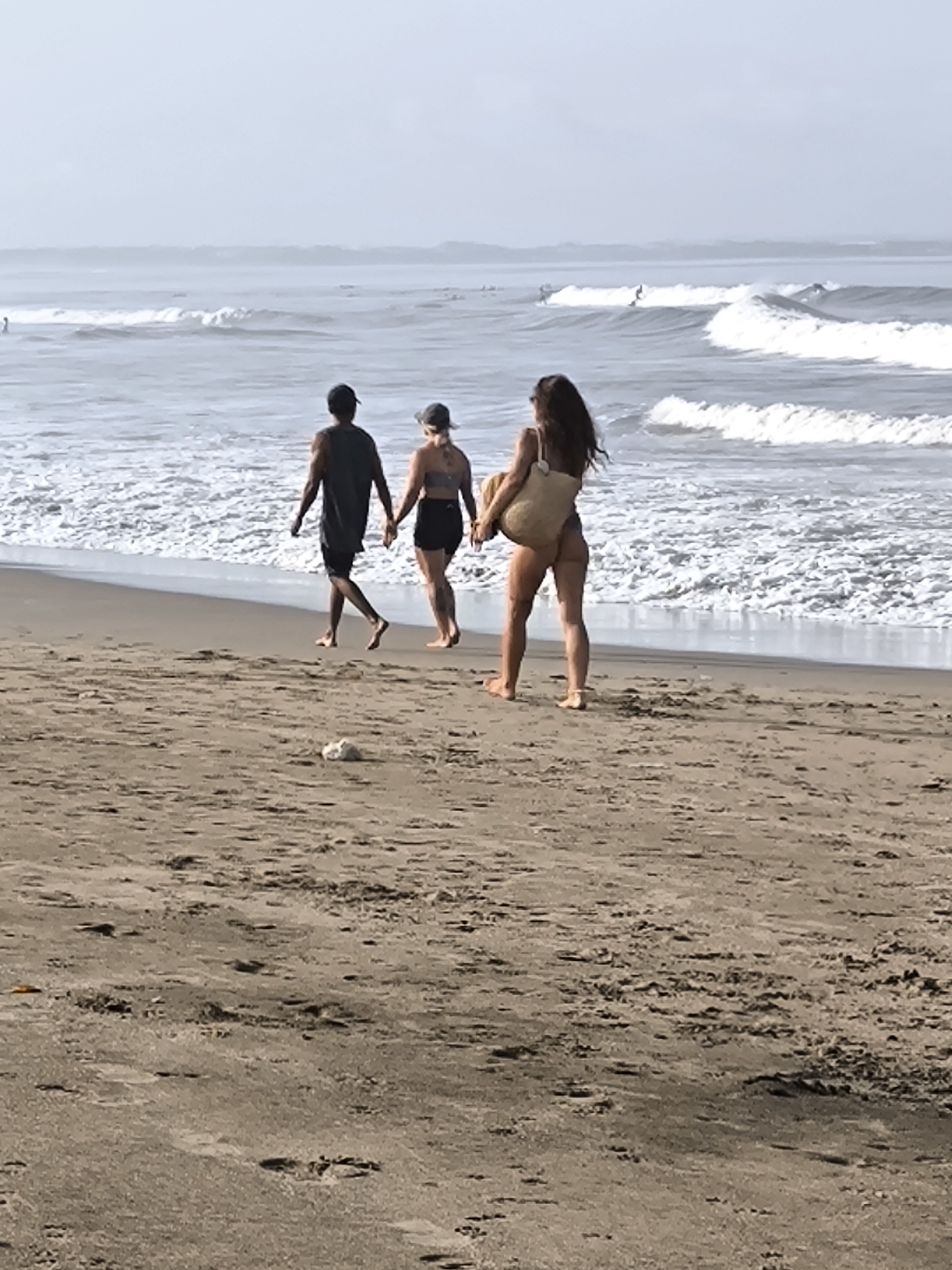 The photograph depicts three individuals walking along a beach in Canggu, Indonesia. A man in a dark shirt and shorts holds hands with a woman wearing a baseball cap, a dark bathing suit top, and dark shorts. A third woman walks slightly ahead, wearing a light-colored bathing suit and carrying a large woven bag. The individuals are barefoot. The beach is sandy, with the ocean visible in the background. White waves are breaking in the distance, where there appear to be some surfers. The sky is overcast.