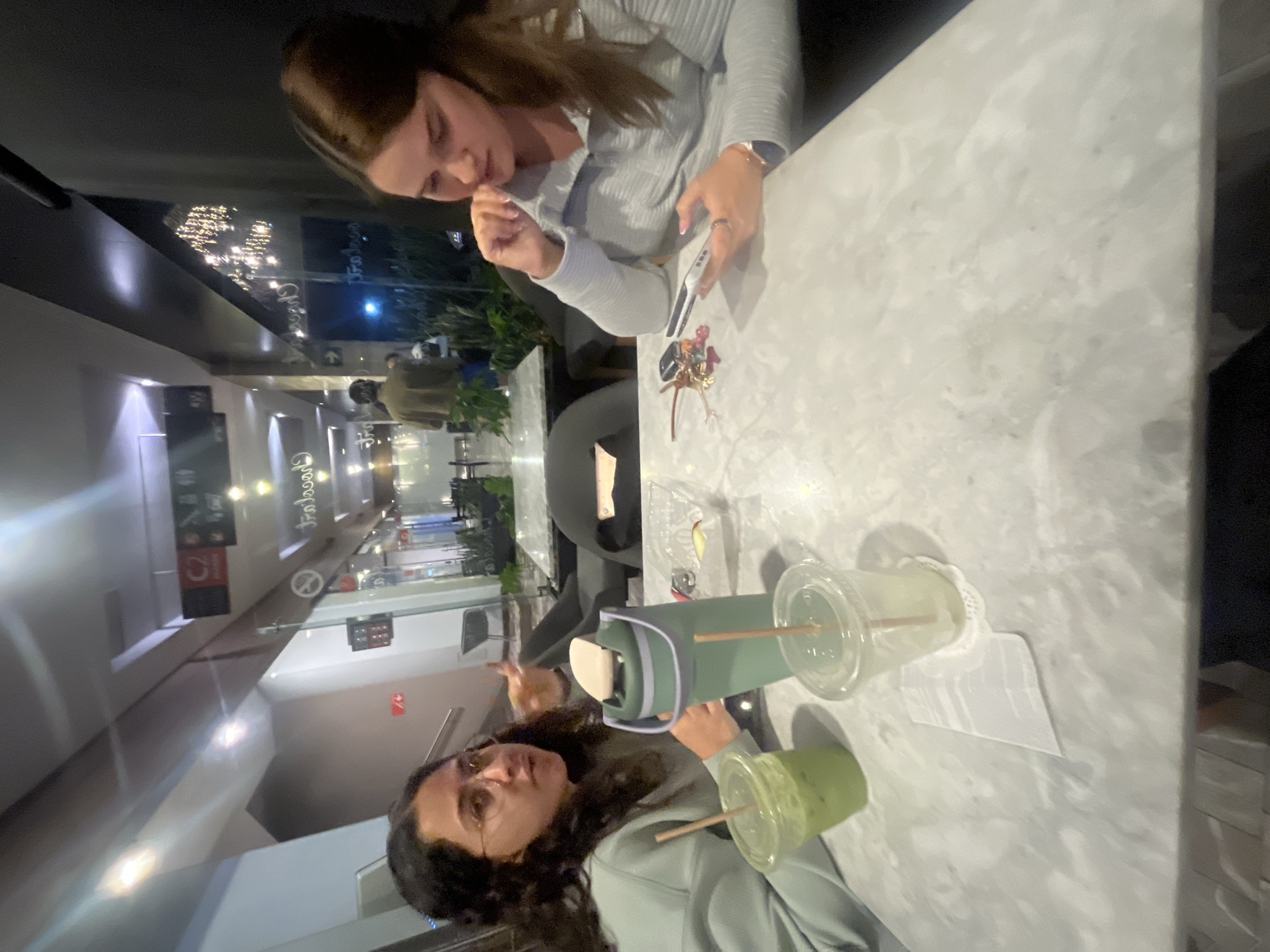 The image depicts two women seated at a light-colored, possibly marble-patterned table within an indoor commercial establishment, identified as a shopping center in the city of Puebla, Mexico. The woman on the left, with dark curly hair and clear-framed glasses, wears a light green long-sleeve top. She is shown in mid-speech, with her mouth open and her right hand slightly raised, gesturing. On the table in front of her are a clear plastic cup containing a green beverage with a straw, and a tall green insulated bottle with a beige lid. A second clear plastic cup, partially empty with a straw, rests on a small white doily next to a folded white napkin. The woman on the right, with long brown hair, wears a light blue ribbed long-sleeve top. She is looking down at a white smartphone held in her hands, with her right hand positioned near her mouth or chin. A set of keys with multiple fobs and charms lies on the table to her left. A clear plastic container holding sliced fruit, likely apples, is situated between the two individuals. In the background, an individual wearing a brown jacket is partially visible, walking away from the camera beyond a glass partition. The environment features glass walls, reflective surfaces, overhead lighting, and signs, including a "C2" indicator with an arrow, a no-smoking symbol, and text that includes "Chocolates" and a reversed "Frascaro" visible on the glass, indicating retail spaces. Potted plants and additional seating areas are also visible in the background.