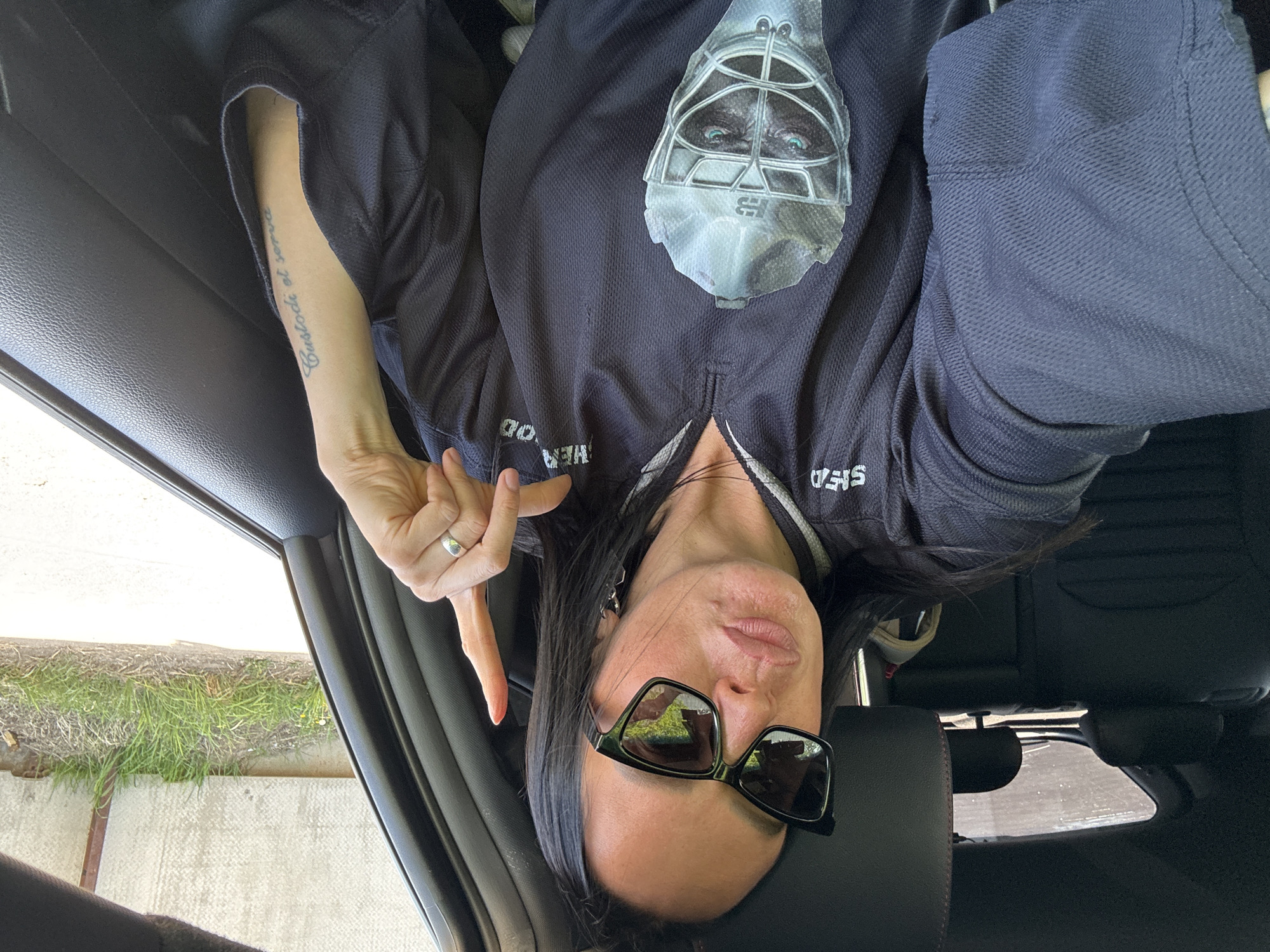 A woman is in a car, wearing a dark jersey with an image of a goalie mask, sunglasses, and a ring on her left hand. She has a tattoo on her left forearm and is making a hand gesture. Outside the car window, there is grass and a concrete surface. There is no indication that this is Chaikovsky, Russia.