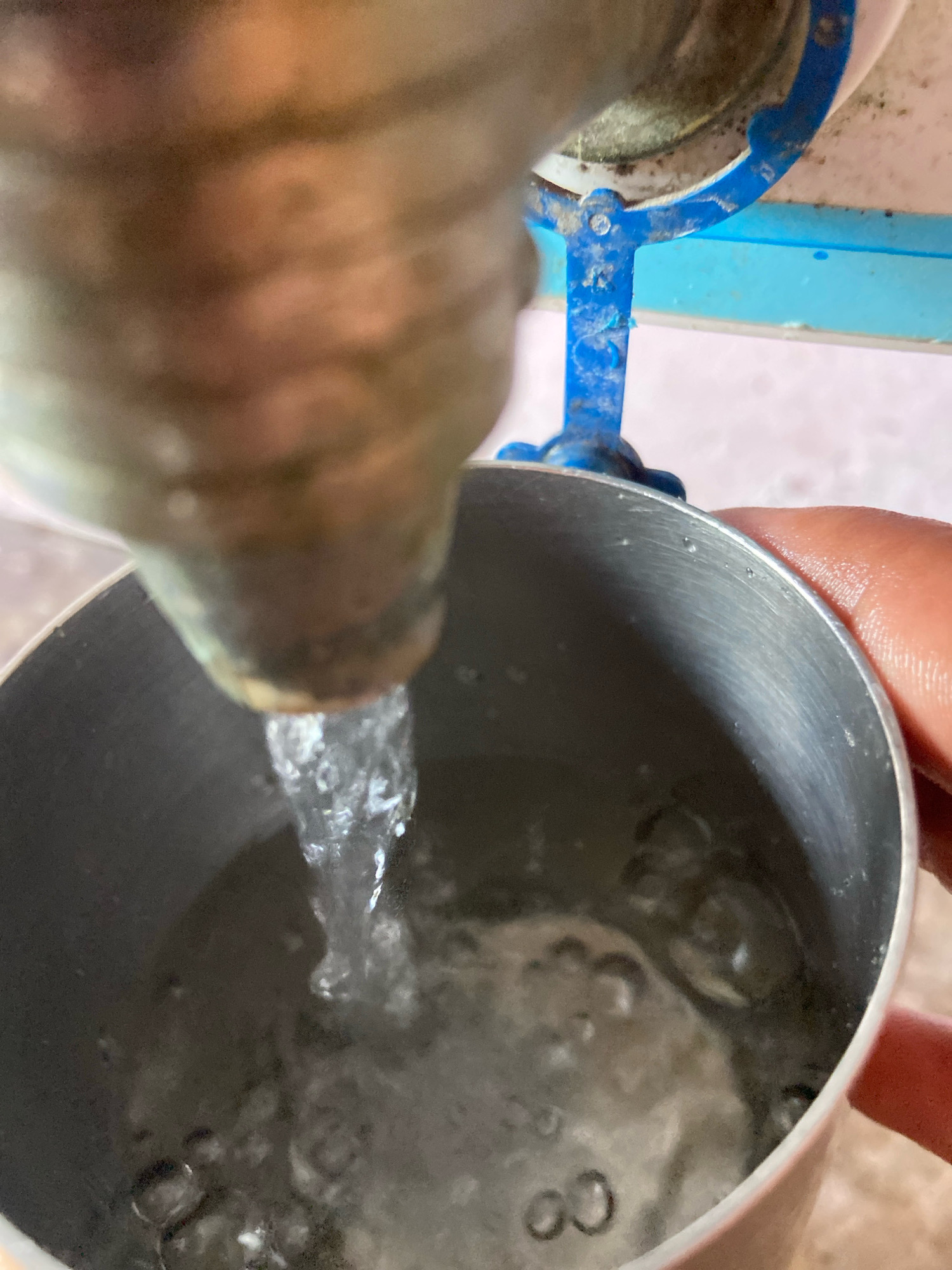 A close-up, shallow depth-of-field shot shows water being poured from a metal faucet into a metal cup. The faucet appears to be made of brass or a similar metal and is mounted on a blue bracket, suggesting it is part of a water dispenser or plumbing fixture. The water stream is clear and appears to be flowing steadily. The metal cup is cylindrical with a brushed metal finish and is held by a person's hand, partially visible on the right side of the frame. The water level in the cup is rising, creating ripples and bubbles on the surface. The background is out of focus but appears to be a light-colored surface, possibly a wall or countertop. The scene is likely taking place in a domestic or public setting where water is being dispensed, such as a home, office, or a public facility. The image does not contain specific visual cues to pinpoint the exact location in Bahadurpur, India, but it depicts a common everyday activity.