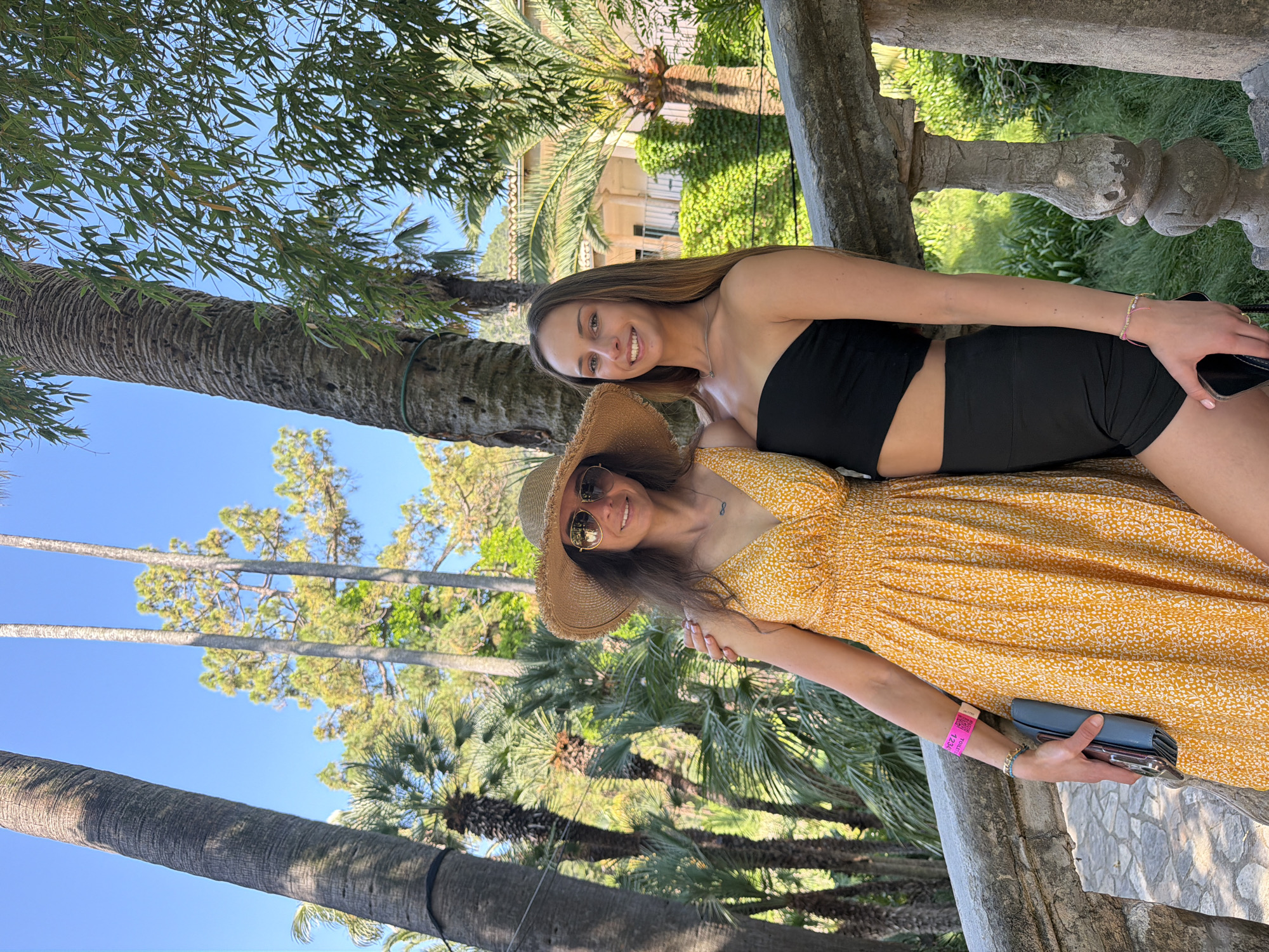 Two women stand close together outdoors. One woman, wearing a yellow patterned dress and a straw hat with sunglasses, holds a cell phone and wallet. The other woman wears a black strapless top and black shorts. They stand next to a stone railing. Palm trees and other greenery are visible in the background. Based on the architecture and landscape, the location appears to be in Bunyola, Spain.