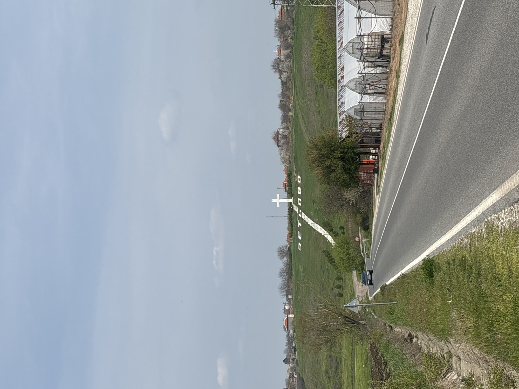 The image presents an outdoor scene in Zetiovo, Bulgaria, featuring a road that runs alongside a grassy field. In the field, a large white cross stands prominently. Adjacent to the road on the right, there are greenhouse-like structures. A car is positioned on the roadside. The sky is overcast.