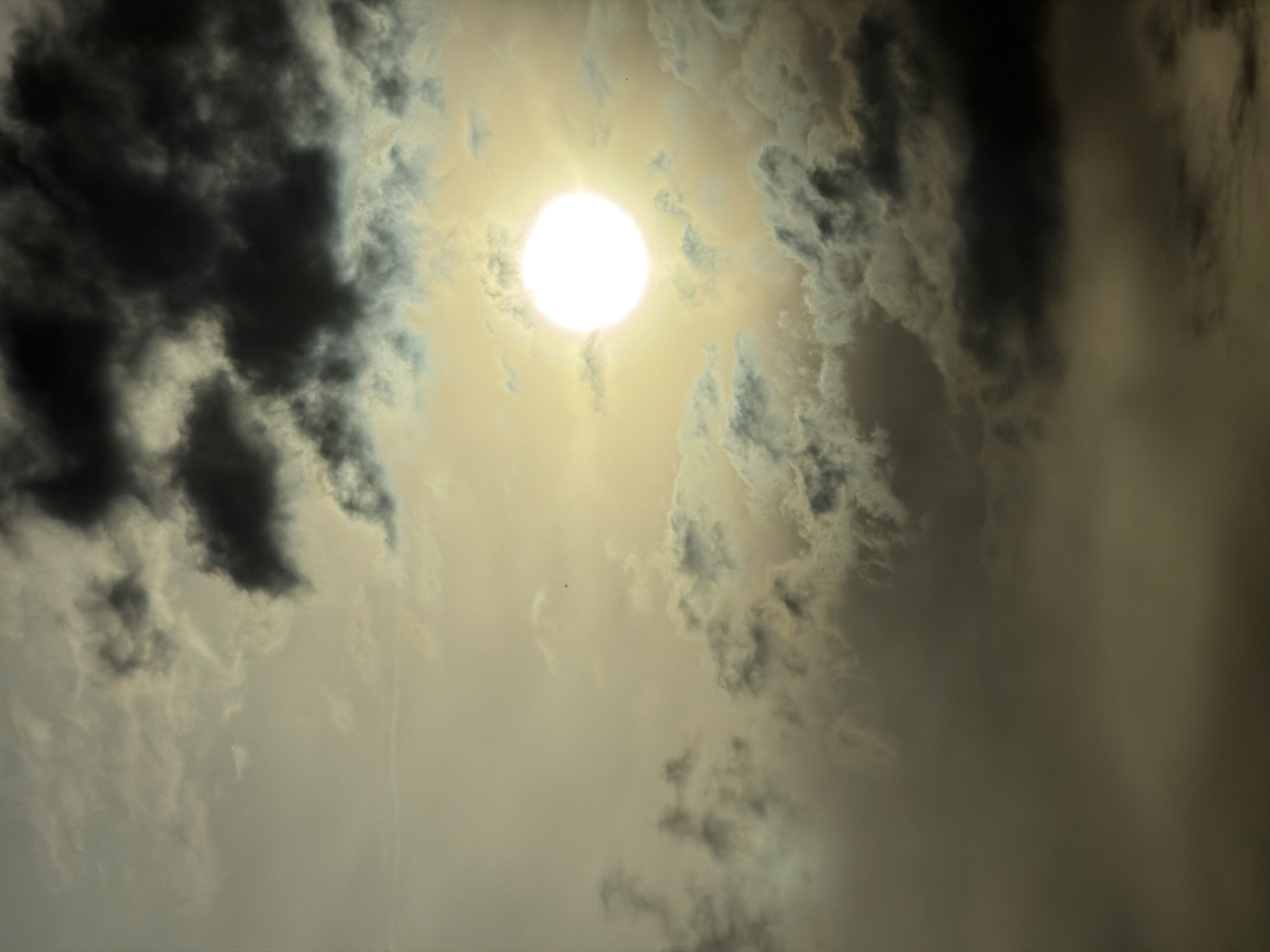 The image depicts a sky filled with dramatic clouds and a bright, diffused sun. Dark, heavy clouds occupy the upper and lower portions of the frame, with lighter, more wispy clouds scattered throughout. The sun appears as a bright, hazy orb in the upper right quadrant, its light creating a glow that illuminates the surrounding clouds. The overall impression is one of a turbulent sky, possibly before or after a storm. There are no identifiable buildings, landmarks, or people in the image, and therefore, no specific location in the city of Mramor, Bulgaria, can be determined.