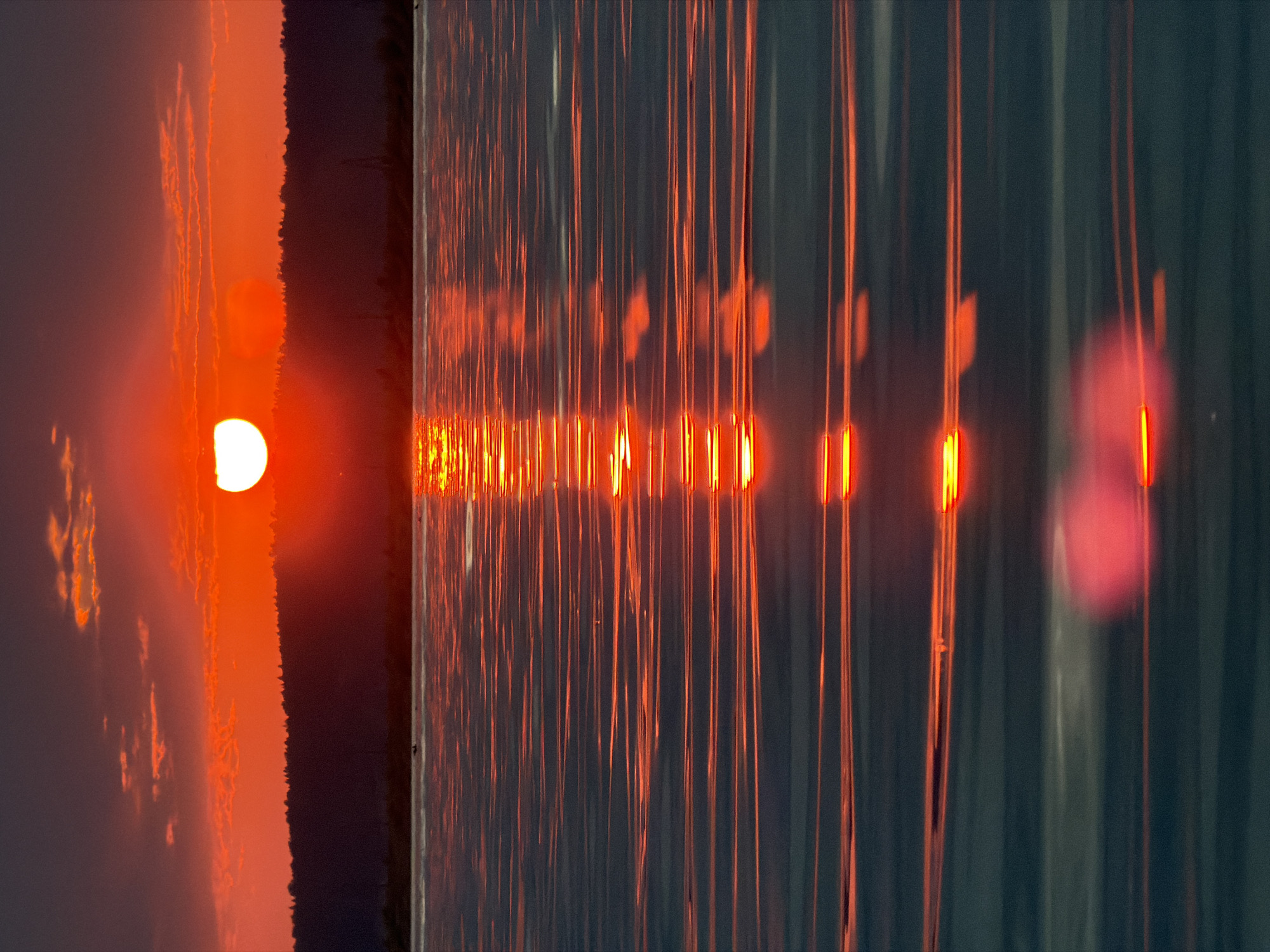 A vibrant sunset casts a fiery orange glow across the sky, illuminating the clouds and reflecting on the surface of a calm body of water. A silhouetted line of trees forms the horizon, beyond which the sky transitions to a deep purple. The reflection of the setting sun creates a shimmering, elongated path of light on the water, broken into horizontal streaks by subtle ripples. The scene appears to be a natural landscape with no discernible urban elements or human presence, suggesting a tranquil, remote location. The overall impression is one of natural beauty and serenity at the end of the day.