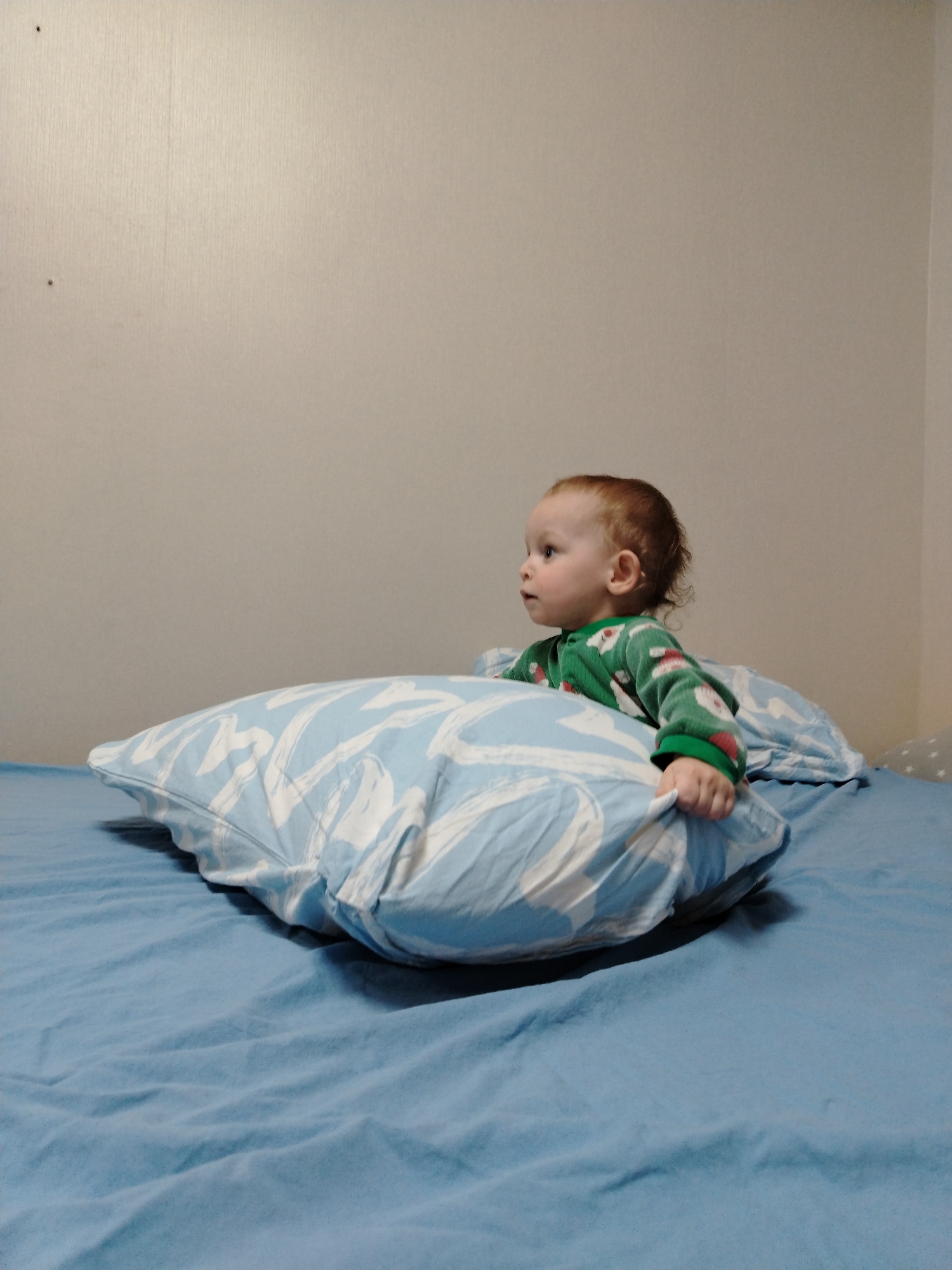 The image displays an infant or toddler positioned on a bed in an indoor setting within Chaikovsky, Russia. The child, exhibiting light reddish-brown hair and fair skin, is dressed in a long-sleeved green top adorned with white and red festive patterns. The child is propped up by a light blue pillow featuring a white abstract design, looking intently towards the left of the frame. The child's left hand is gripping the patterned pillow. The bed is covered with plain light blue sheets. A second, partially visible pillow with a faint pattern is situated behind the child on the right. The background is a plain light beige wall, which contains two small, dark, circular marks in the upper left area and a visible interior wall corner on the far right.