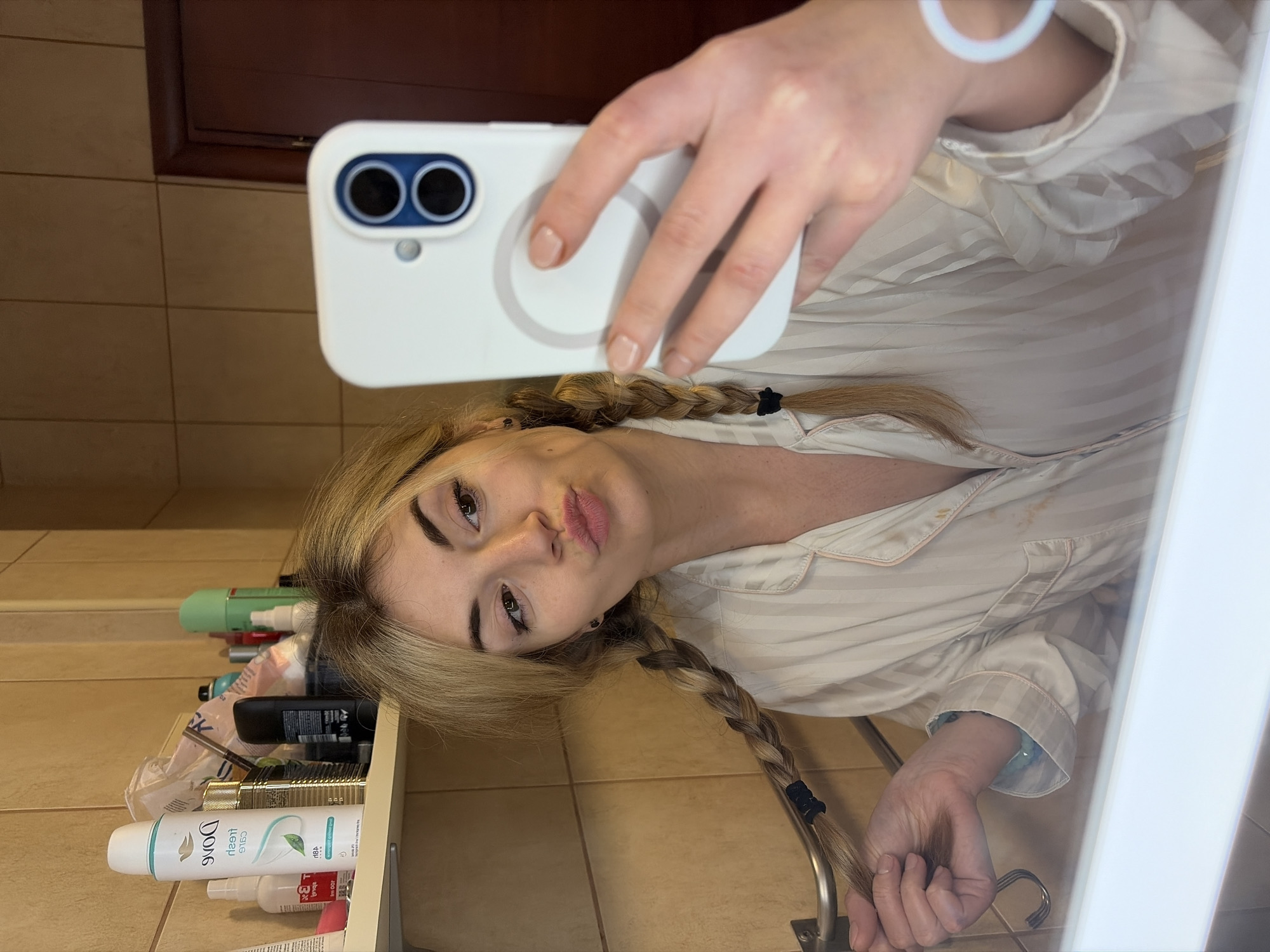 The image depicts the reflection of a female subject in a bathroom mirror. The subject has blonde hair styled in two braids, each secured with a black elastic band. She wears a light-colored, vertically striped collared top with pink piping. Her lips are pursed in a pout, and she has visible eyeliner. She holds a white smartphone, encased in a white case featuring a circular magnetic charging ring and a dual-lens camera module with blue accents, in her right hand. Her left hand loosely holds the end of her left braid, which is draped over a metallic horizontal bar. On her visible right wrist is a light blue-green bracelet, and on her left wrist, a thin white circular bracelet. Above the subject's head, a white shelf holds various toiletries, including an inverted white Dove "Fresh Care" deodorant can, a gold-colored cylindrical bottle, and other personal care products. The background features light brown tiled walls and a partial view of a dark brown wooden door frame. A white basin or bathtub rim is visible in the lower foreground. The exact location within the city of Budva, Montenegro, is not discernible from the provided image, but the setting is a private bathroom.