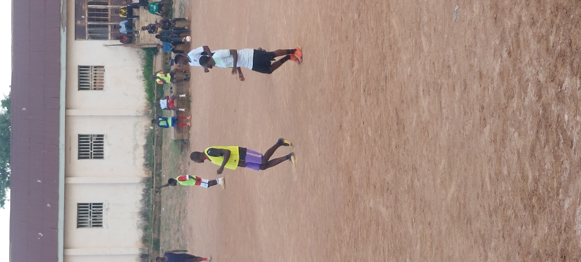 In Bukuru, Nigeria, a group of young men are engaged in a soccer training session on a dirt field. The men are wearing athletic attire, including jerseys, shorts, and cleats. Some are actively participating in drills, while others observe from the sidelines. In the background, a light-colored building with a corrugated metal roof and barred windows stands behind the field. The sky is overcast, and the overall atmosphere suggests a typical afternoon of sports activity in a community setting.