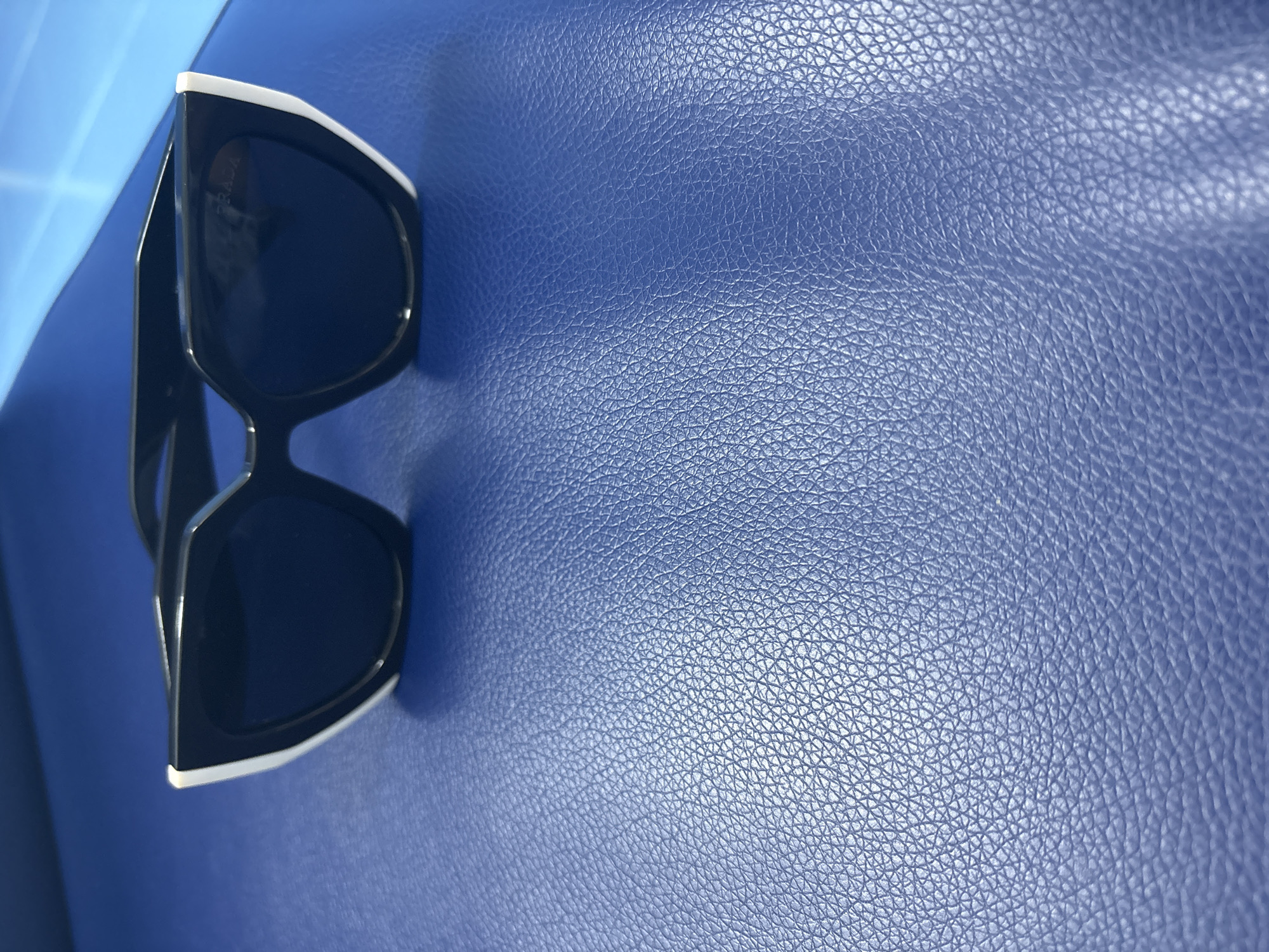 A pair of black-lensed sunglasses with thick white rims rests on a textured blue surface. The sunglasses are positioned on the left side of the frame, angled diagonally from the top left corner towards the bottom right. The blue surface appears to be leather or a similar material, characterized by a consistent, fine-grained texture. The lighting suggests it is daytime, with a bright light source casting subtle shadows and highlighting the texture of the blue surface. There are no people visible in the image. The scene is simple and focused on the object and its immediate surroundings. Some text is faintly visible on the inside of the sunglasses, appearing to be "PRADA". The context of Orsk, Russia, and any specific time of day or weather conditions are not directly inferable from the visual information in the image alone.