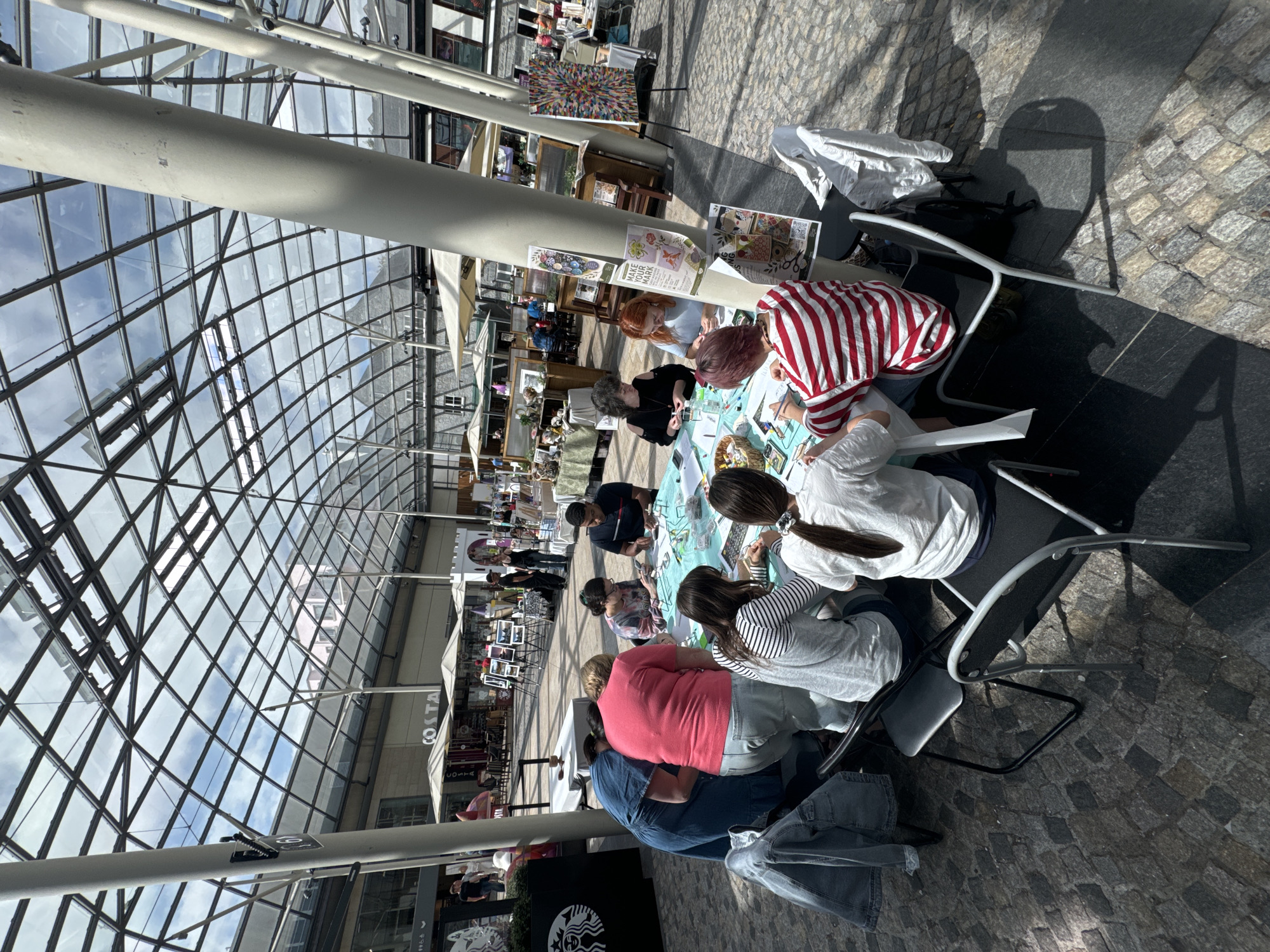 The image shows a group of people gathered around a table, engaged in a crafting activity under a large, glass-covered structure. The scene appears to be an indoor market or public space with a cobblestone floor. Several individuals are seated or standing around a circular table covered with a light-colored cloth. The table is laden with various craft supplies, including beads, scissors, and what appear to be kits or small containers.
In the foreground, on the left side of the image, a woman wearing a denim dress is leaning over the table, looking at the supplies. Next to her, a woman in a pink t-shirt and jeans is also leaning in. To her right, a woman with dark hair and glasses is seated, working on a craft. Further to the right, a man in a dark t-shirt is seated, also engaged with the materials on the table. In the mid-ground, a woman with blonde hair in a grey and white striped shirt is seated, as is a woman in a white t-shirt with her back to the camera, her dark hair in a ponytail. On the far right, a person wearing a red and white striped t-shirt is seated, working on the crafts.
The environment features a large, multi-paneled glass roof that allows natural light to filter through, illuminating the space. Structural supports, including large white pillars, are visible throughout the scene. In the background, glimpses of shops and stalls can be seen, with umbrellas providing shade for outdoor seating areas. There are also posters and artwork displayed on a pole in the mid-ground. The overall impression is of a lively, communal creative session taking place in a well-lit, open-plan venue. This location is the Market Yard in Kilkenny, Ireland, a popular spot for events and markets, covered by a distinctive geodesic dome.